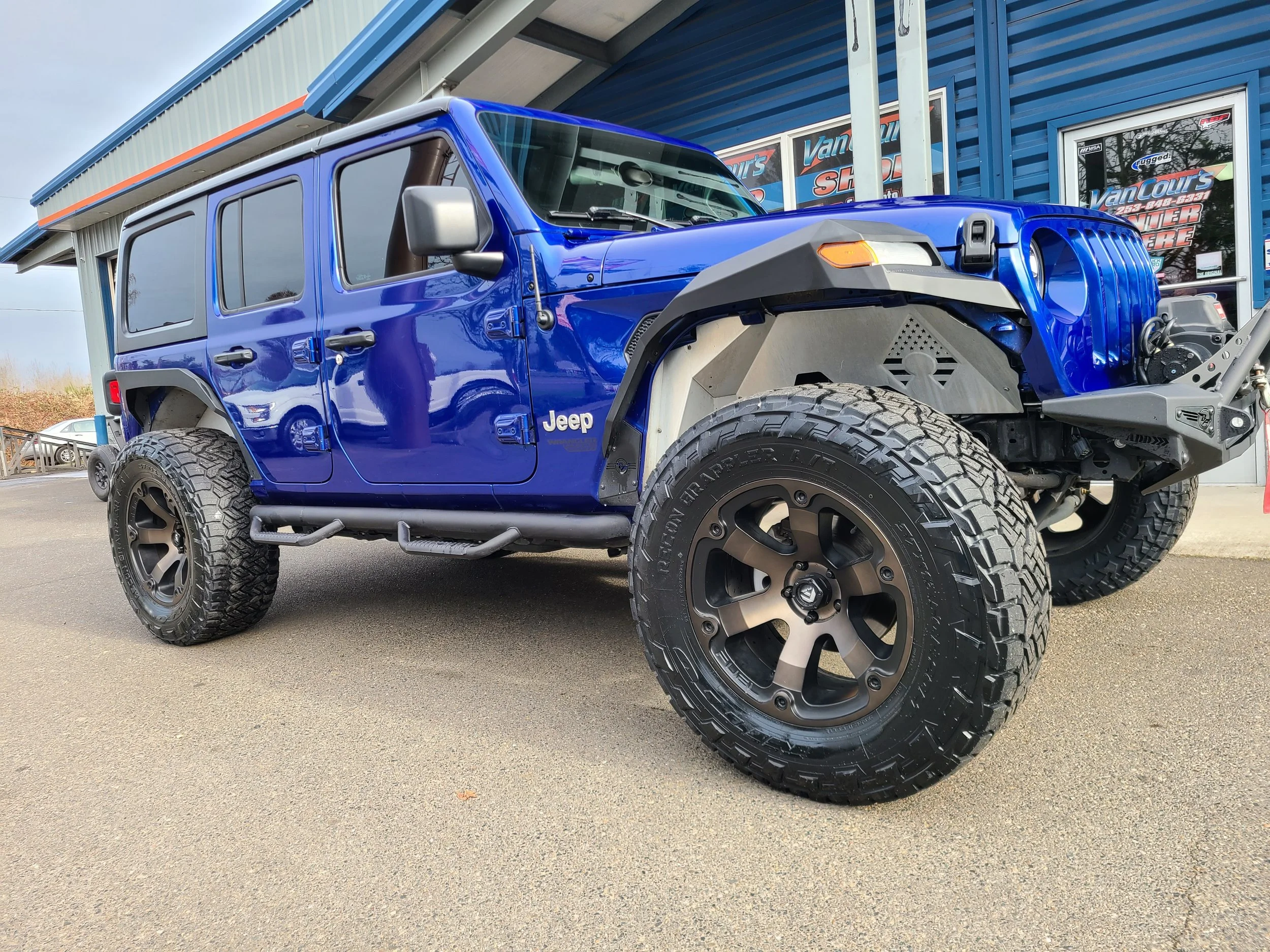 A blue Jeep vehicle with large off-road tires parked outside a building with signs for VanCourt's and Power Store.