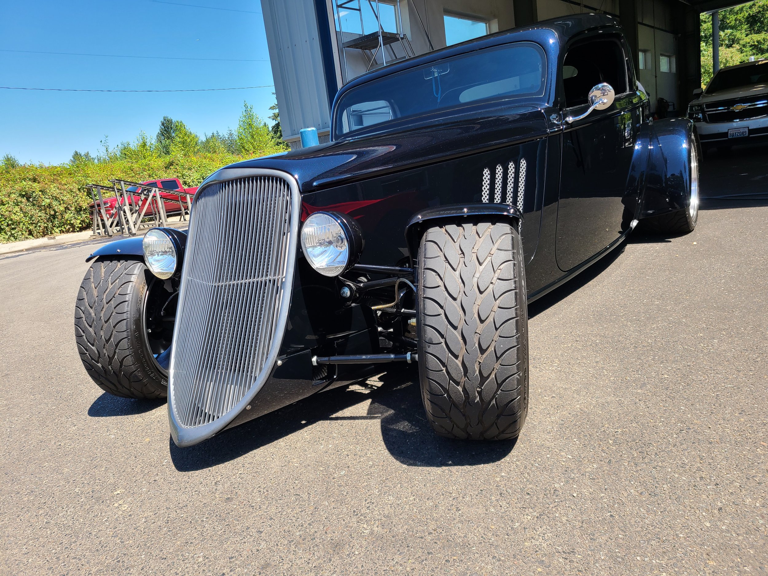 A black vintage car with large tires and a chrome front grille parked indoors. There is another vehicle in the background to the right and some outdoor elements are visible through the windows.