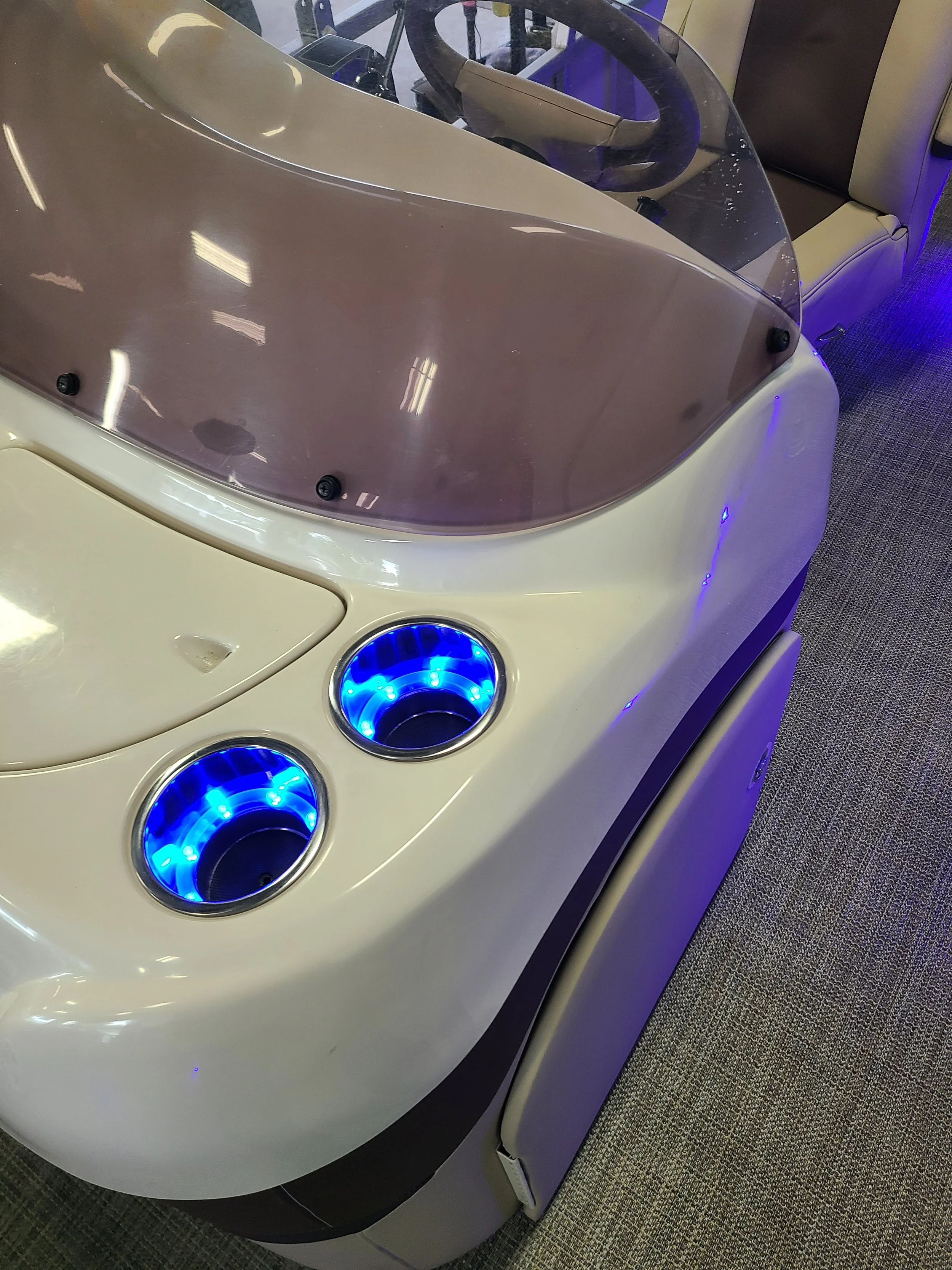 A futuristic simulator with a transparent windshield, blue illuminated cup holders, and a beige seat, situated on a patterned carpet.