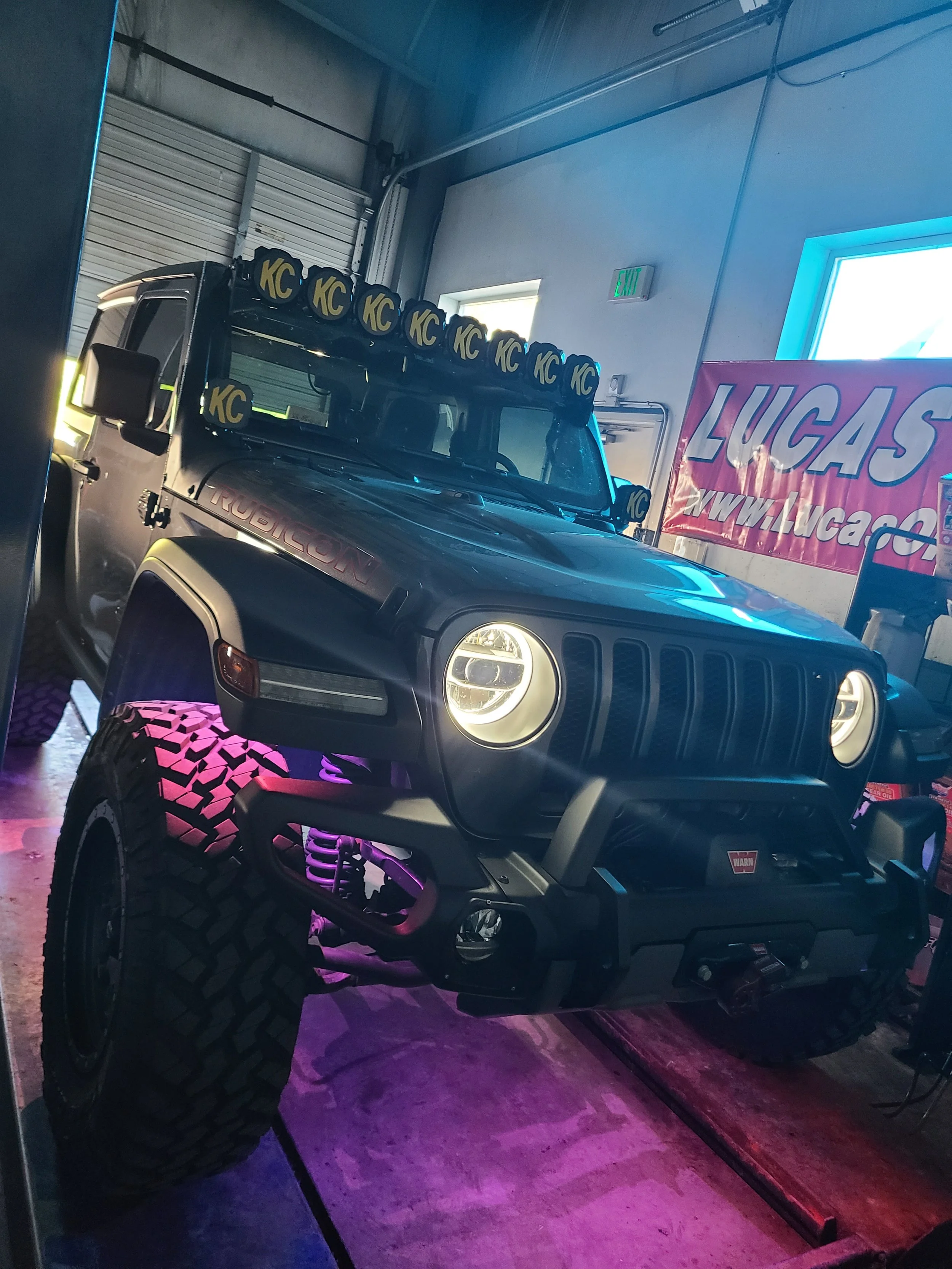A black Jeep Rubicon with large off-road tires and pink accents on the wheels is inside a garage. The vehicle is equipped with KC lights mounted on the roof. There is a pink and purple illuminated sign that reads 'LUCAS' hanging on the wall.