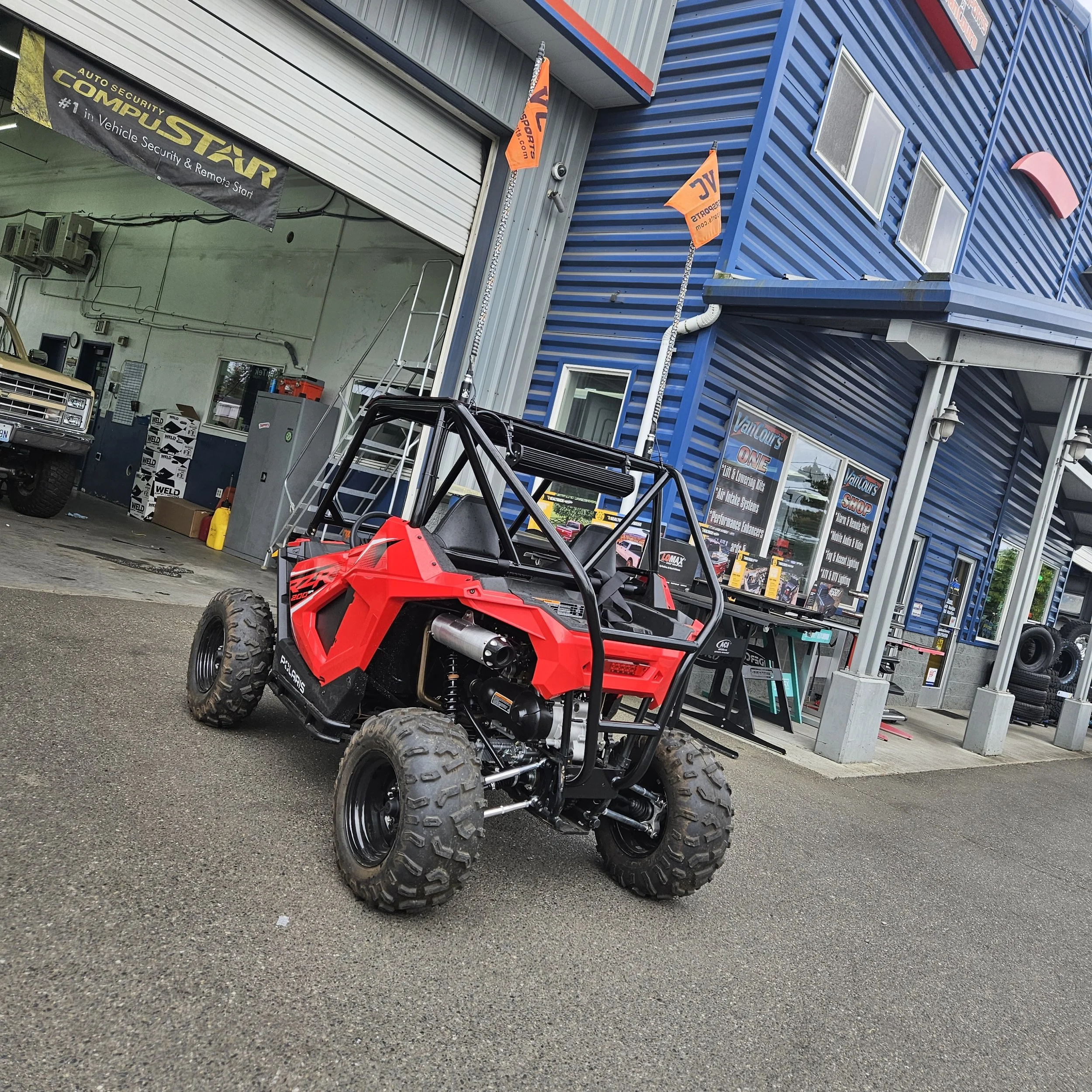 A red off-road vehicle with large tires parked outside a blue building with windows. The building has signage and orange flags, and there are various tools and equipment nearby, including boxes, a yellow container, and a truck.