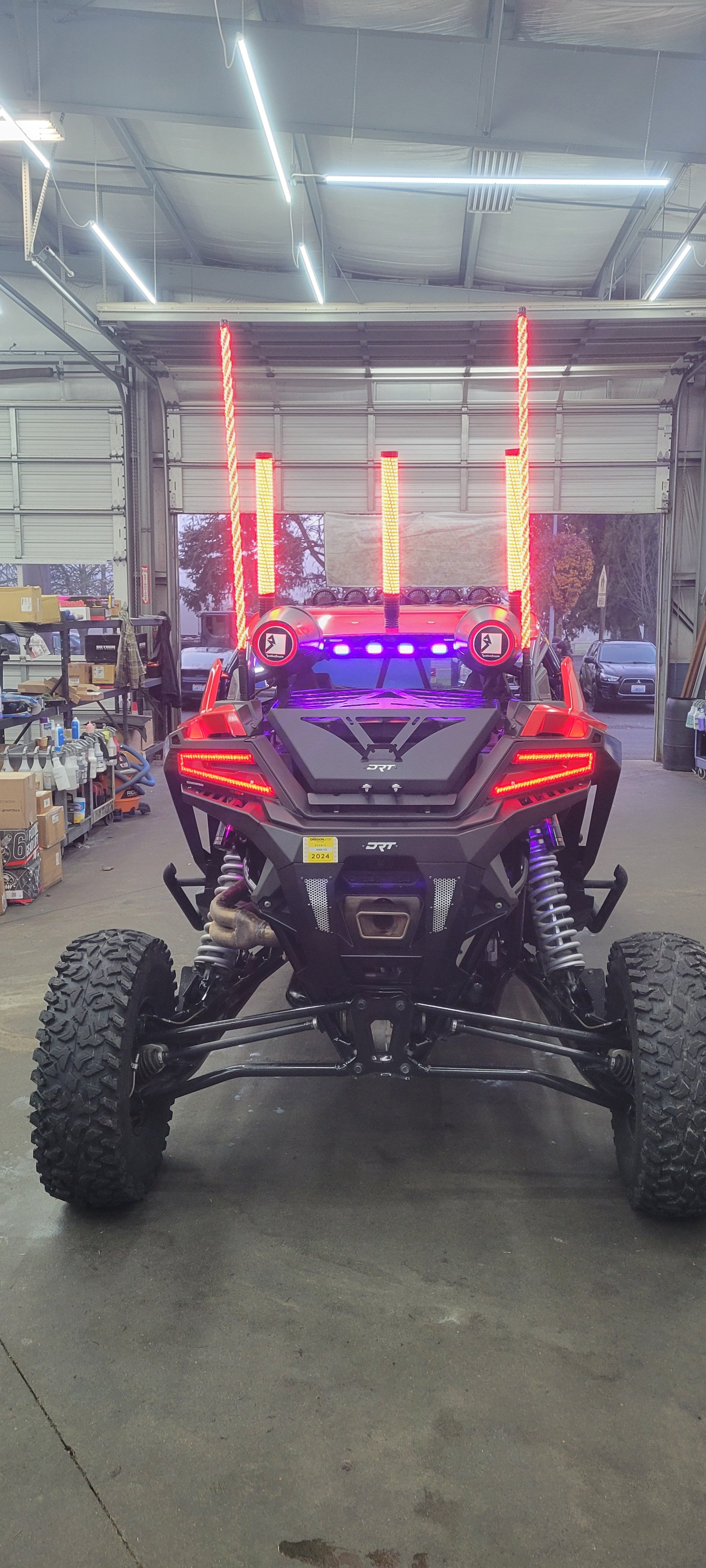 Front view of a black off-road vehicle with large tires, prominent suspension, and LED light bars, parked inside a garage with shelves, tools, and other vehicles visible in the background.
