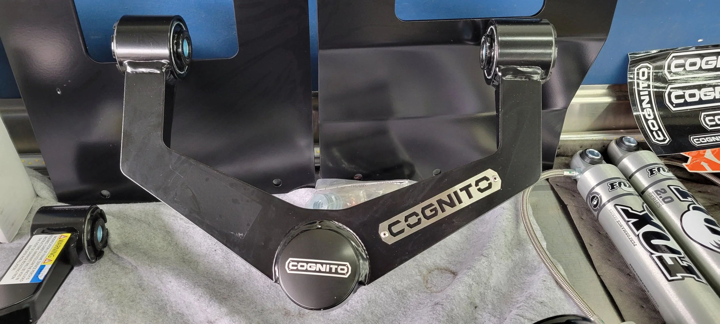 Disassembled motorcycle or vehicle parts, including a black metal frame labeled 'COGNITO', several shock absorbers labeled 'FOX 2.0' and 'FOX 2.5', and other small components on a work surface.