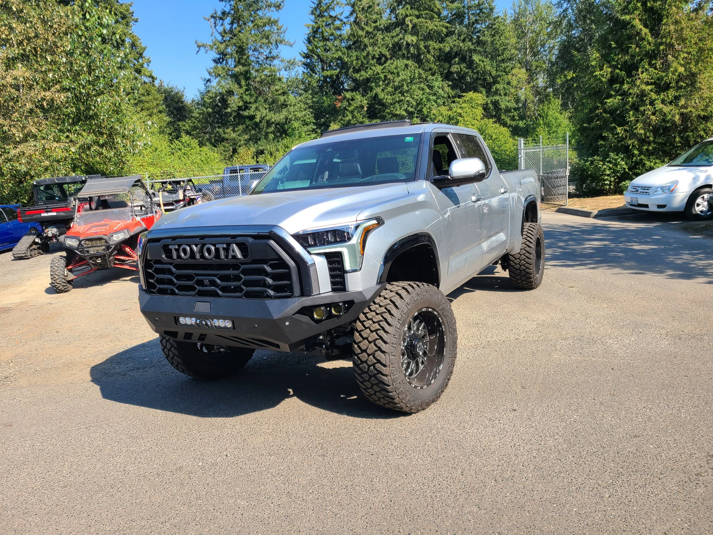 A silver Toyota pickup truck parked outdoors in a lot with trees in the background, alongside other vehicles including a white sedan and a red and black off-road vehicle.