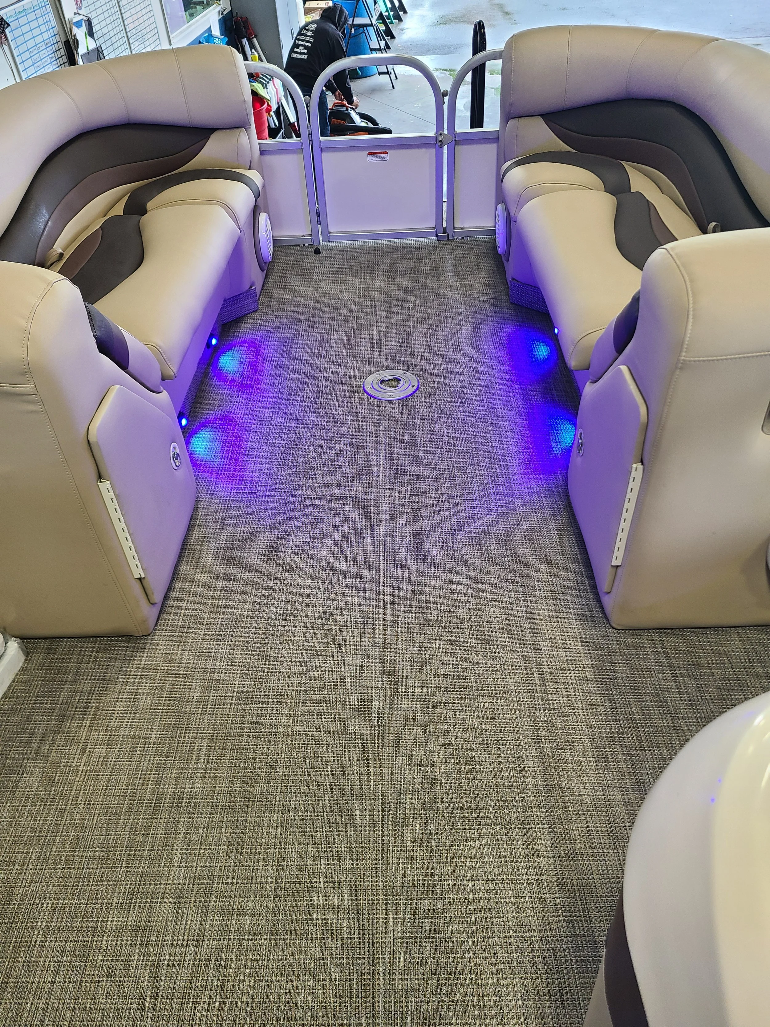 Inside a vehicle with beige and dark gray cushioned seats arranged in a semi-circle, illuminated with blue LED lights on the floor, and a metal barrier at the front, with a person working outside near a blue chair.