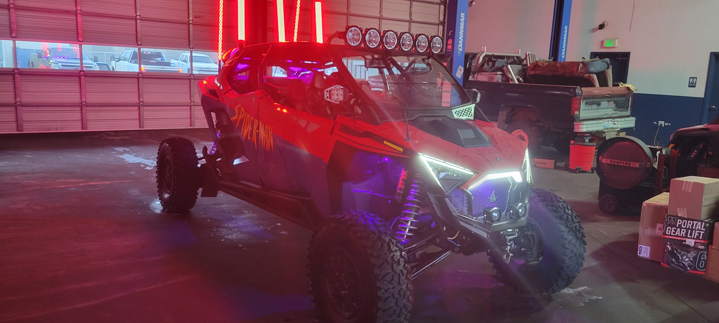 Red off-road vehicle with 'Spiderman' written on the side, equipped with multiple lights on top, in a garage with various tools, equipment, and a closed garage door visible in the background.