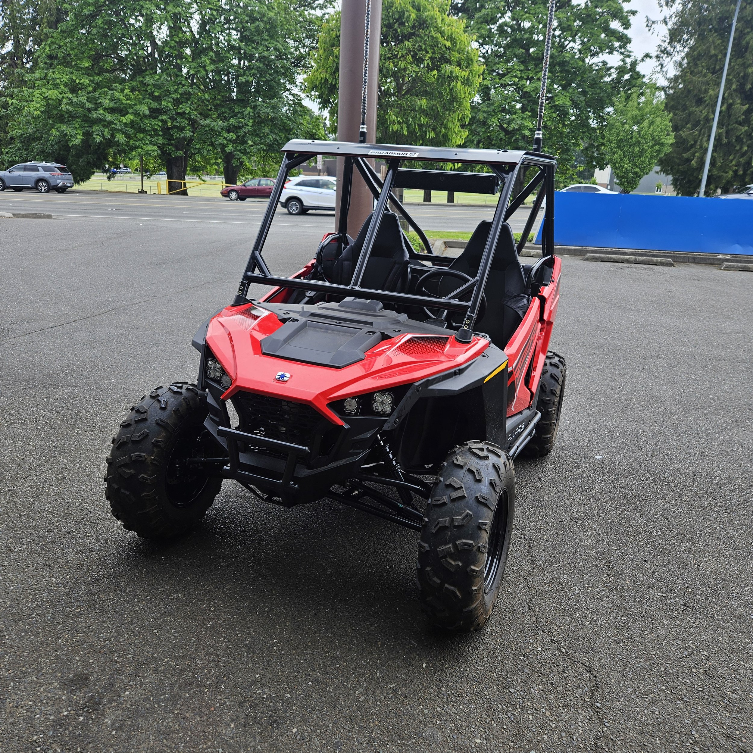 Red and black off-road utility vehicle parked on a paved lot with green trees in the background.