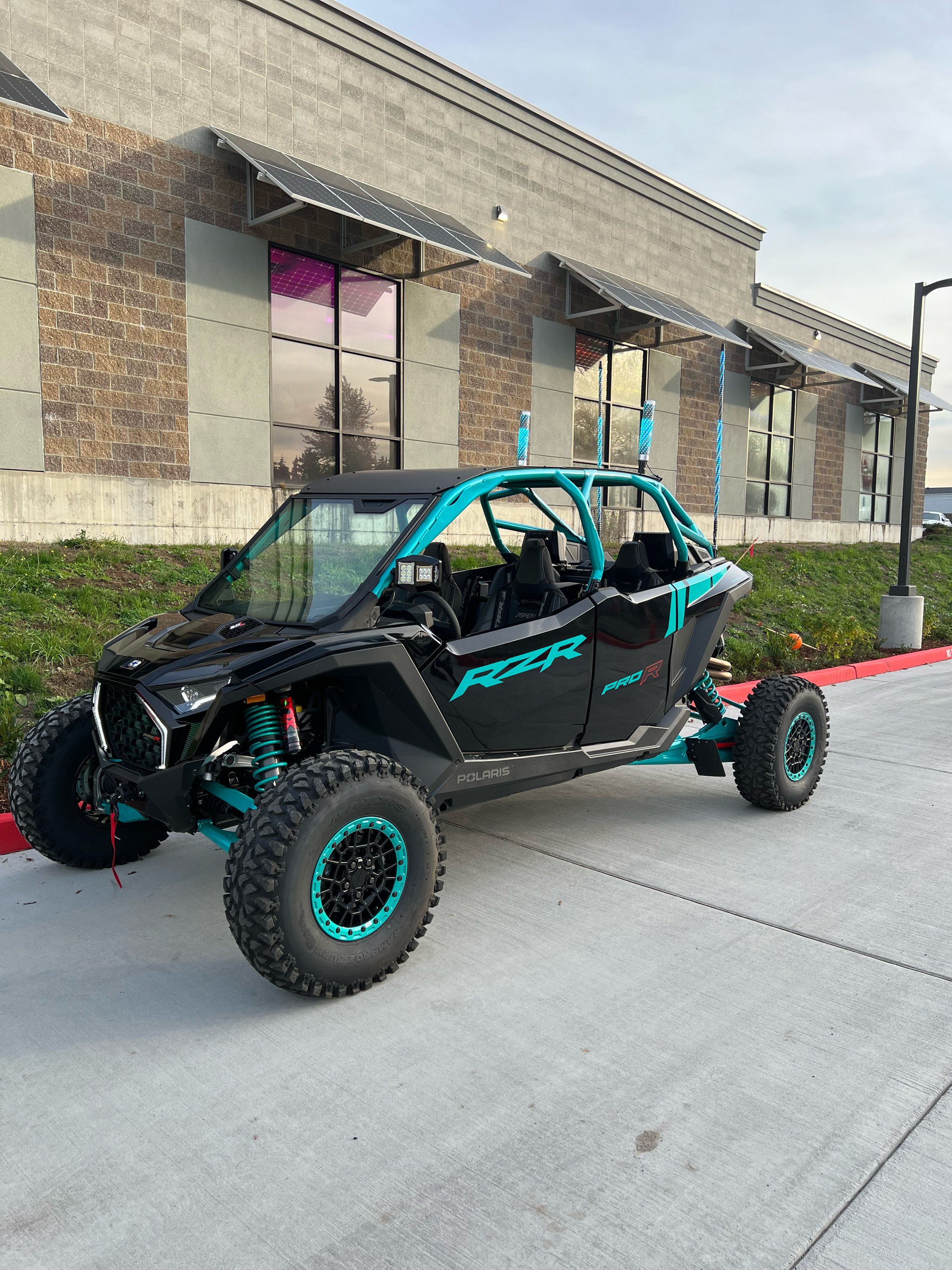 Black and teal Polaris RZR off-road vehicle parked outside near a building with solar panels and tinted windows.