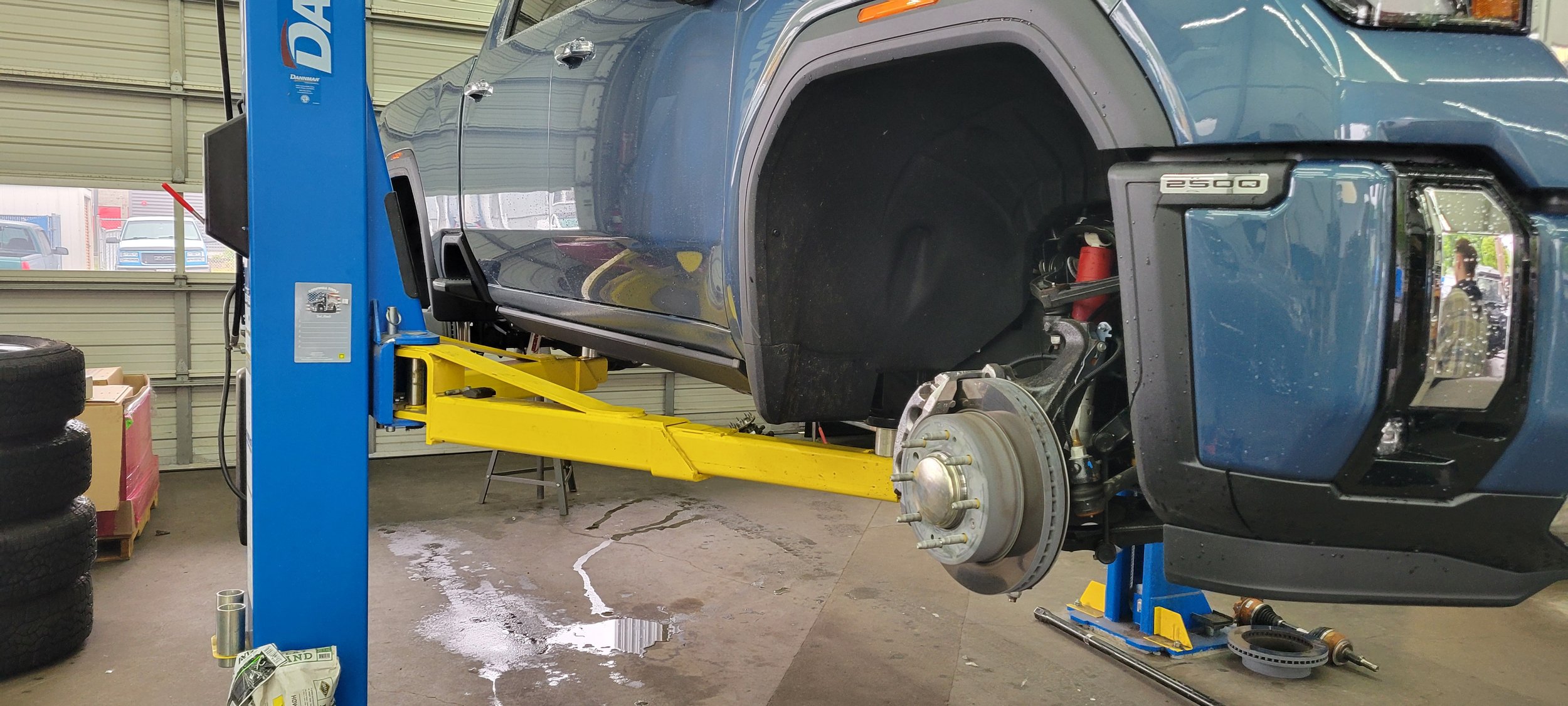 Uncar with no front wheels raised on a hydraulic lift inside an auto repair shop, with tools and spare tires nearby.