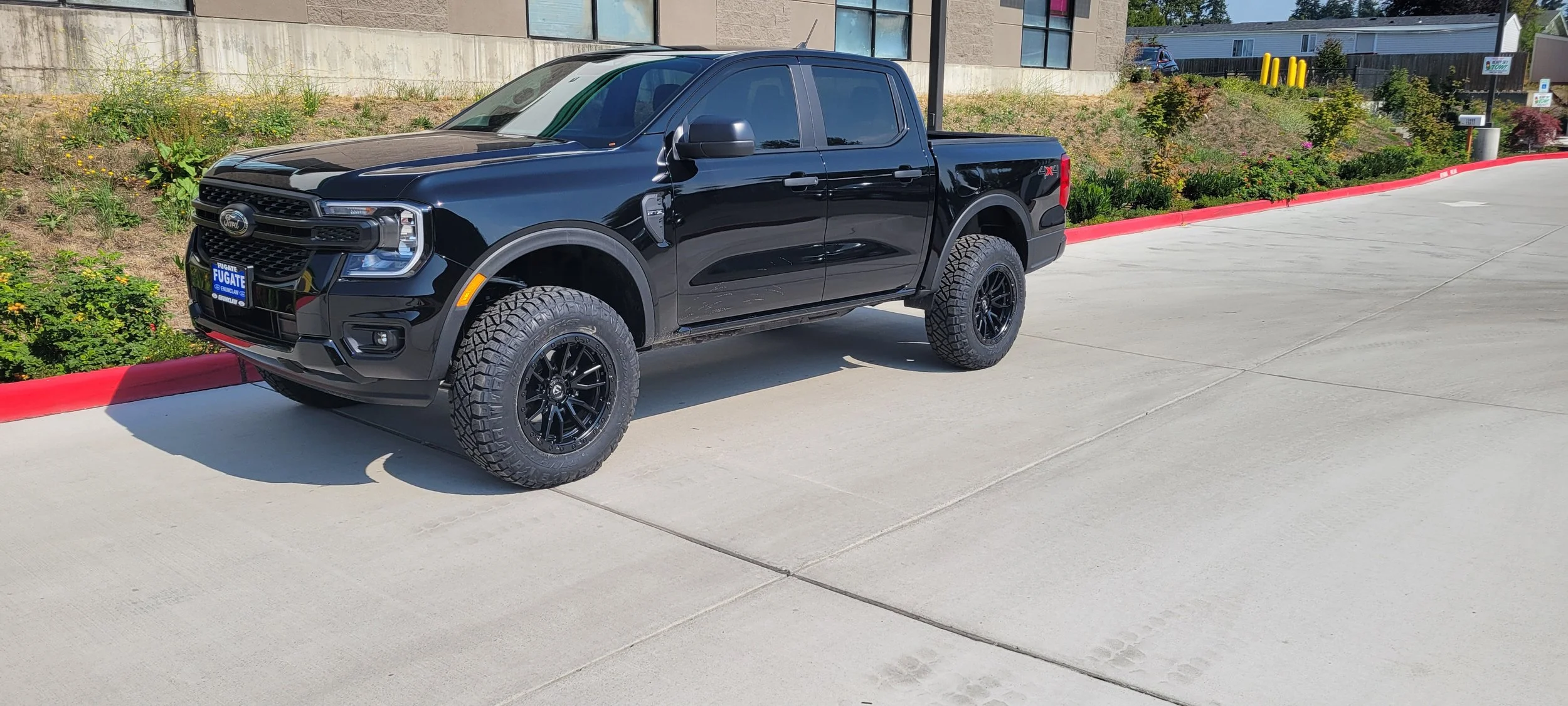 A black Ford pickup truck with four doors and off-road tires parked on a concrete surface near a red painted curb in an outdoor area with greenery and buildings in the background.