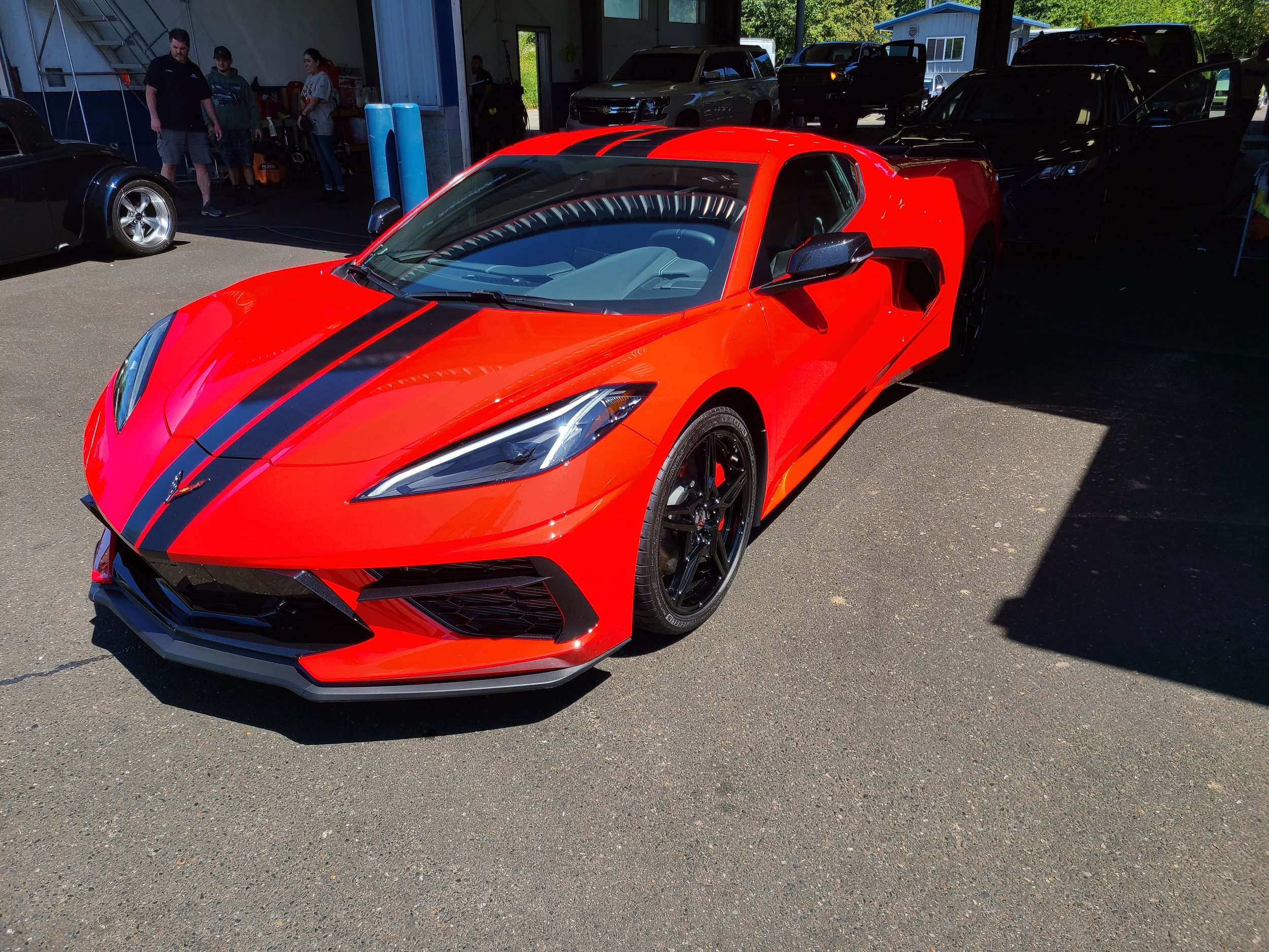 A red sports car with black racing stripes parked inside a garage, with other vehicles and people visible in the background.