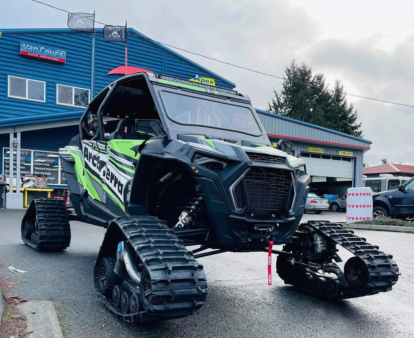 Off-road vehicle with a black and green design and rubber tracks instead of wheels, parked outside an auto shop with a blue building, garage doors, and various cars.