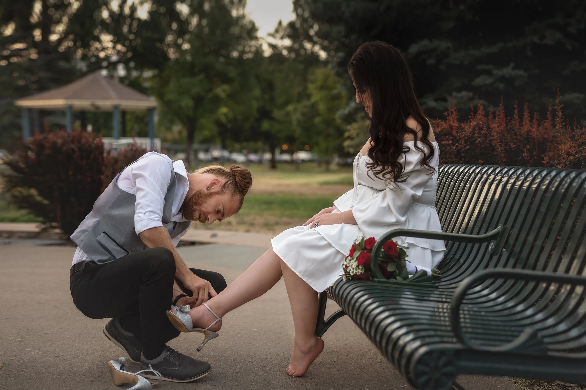 The groom kneeling and fastening the shoe of the bride sitting on a park bench. The bride is wearing a white dress, and a bouquet of red roses is on the bench next to her. Intimate storytelling photography. Wedding and elopement photography. Colorado