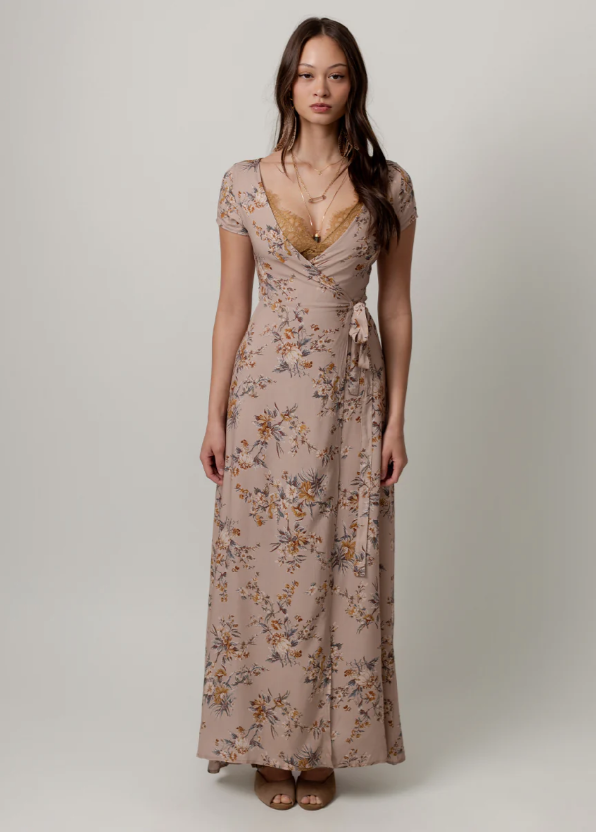 A woman wearing a long, floral dress with short sleeves and a wrap-around tie at the waist, standing against a plain light gray background.