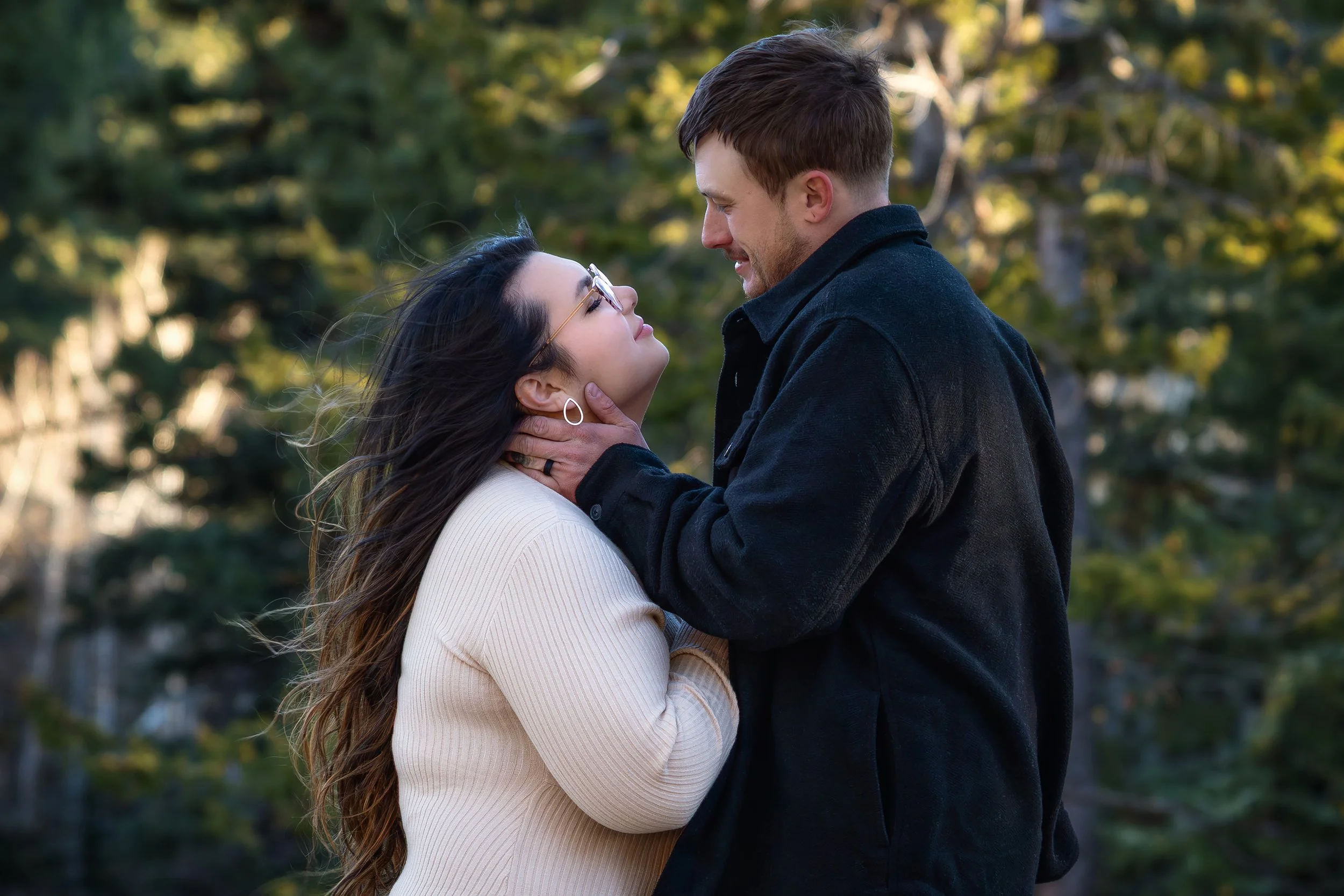 Colorado engagement photoshoot. Colorado engagement photographer. Estes Park Colorado storytelling photographer. Wordlessly, they share an intimate moment, surrounded by blurred trees and foliage in the background. Windblown hair. Colorado forest.