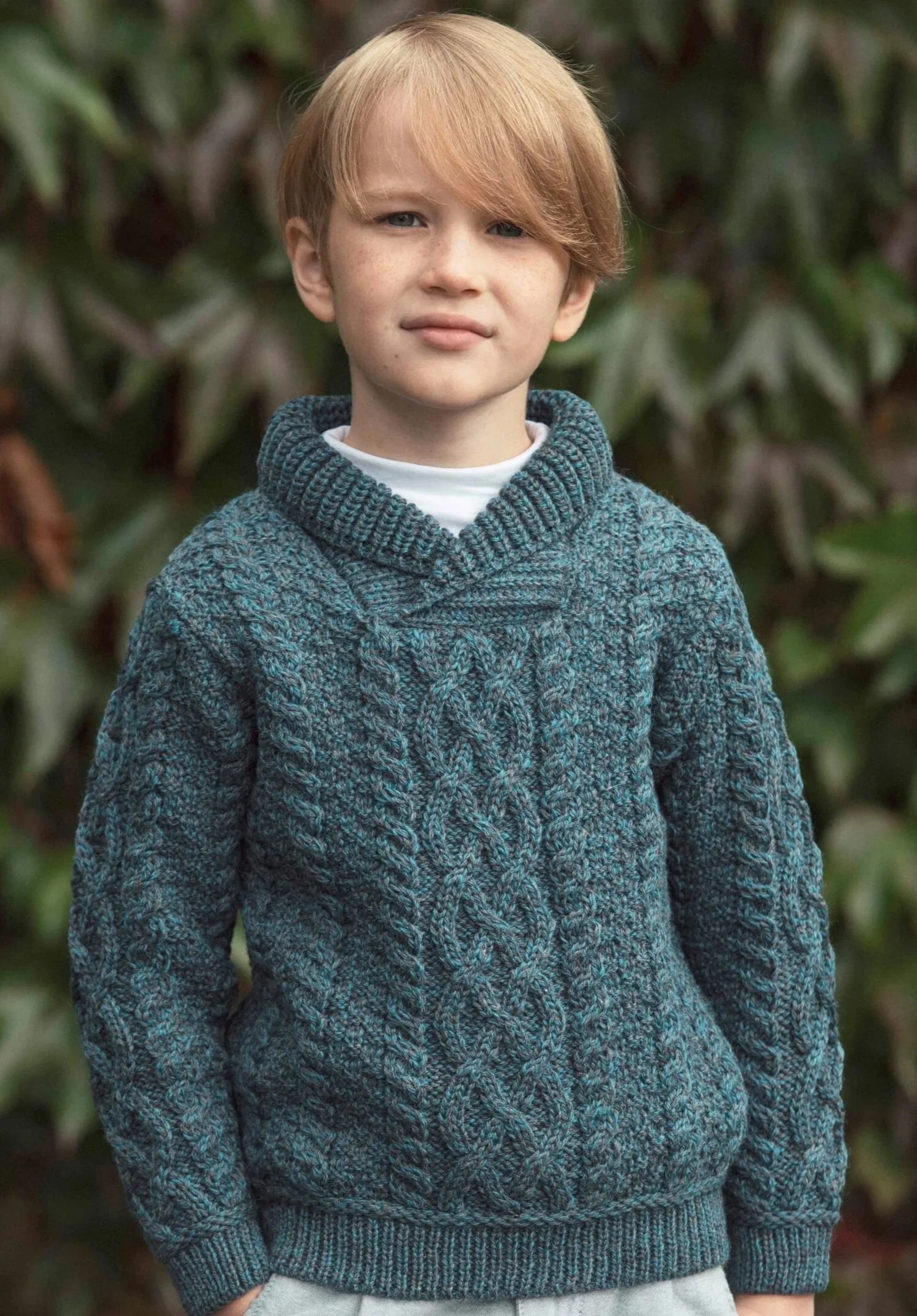 A young boy with red hair and fair skin standing outdoors with greenery in the background, wearing a blue knitted sweater and a white shirt.