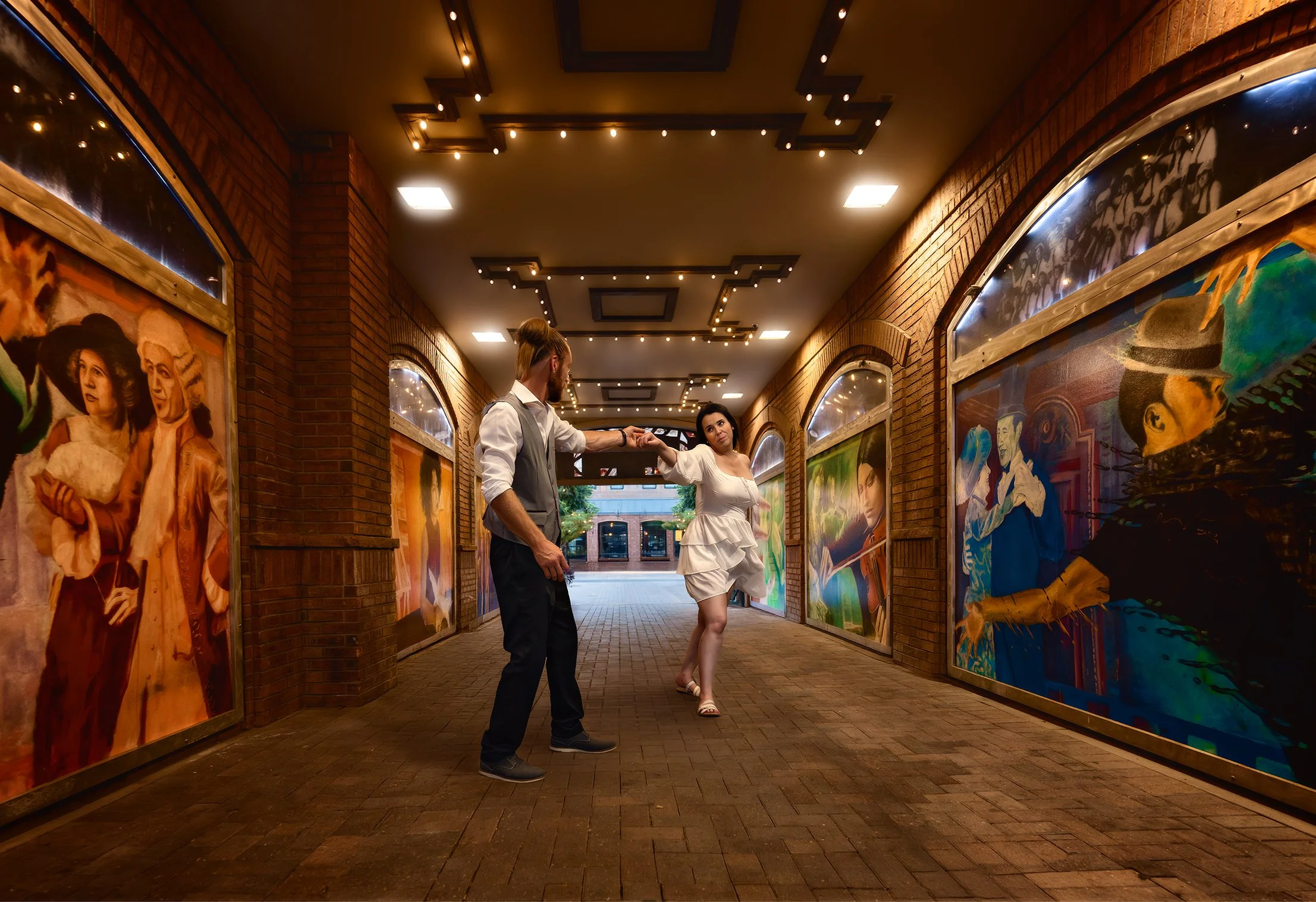A man and woman are dancing together in an art gallery with colorful paintings on brick walls and string lights on the ceiling. Tunnel of murals in Greeley Colorado. Newly weds elopement photoshoot in Colorado
