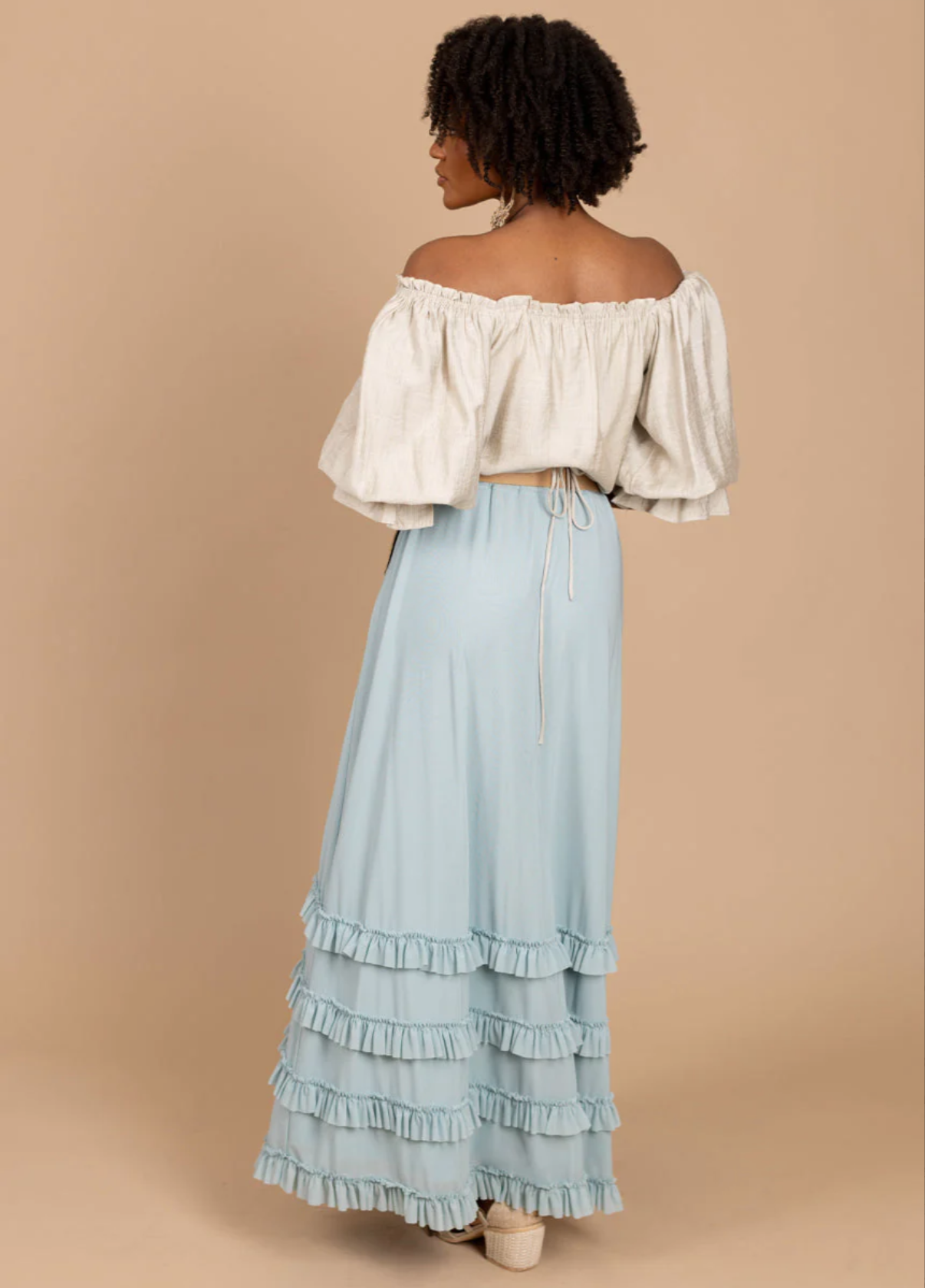 A woman with curly hair wearing an off-the-shoulder beige top and a long, light blue ruffled skirt.