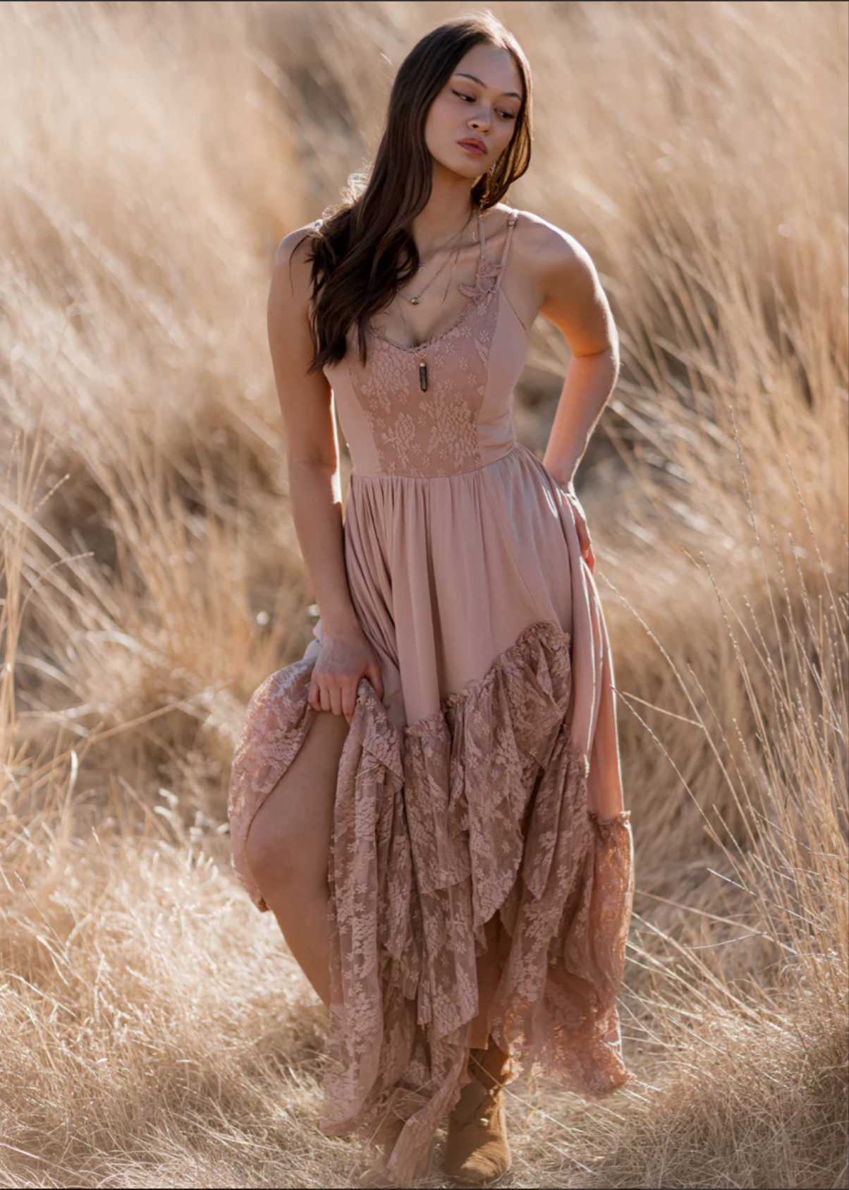 A woman in a pink lace and chiffon dress standing in a field of tall, golden grass, holding up part of her dress while looking down.