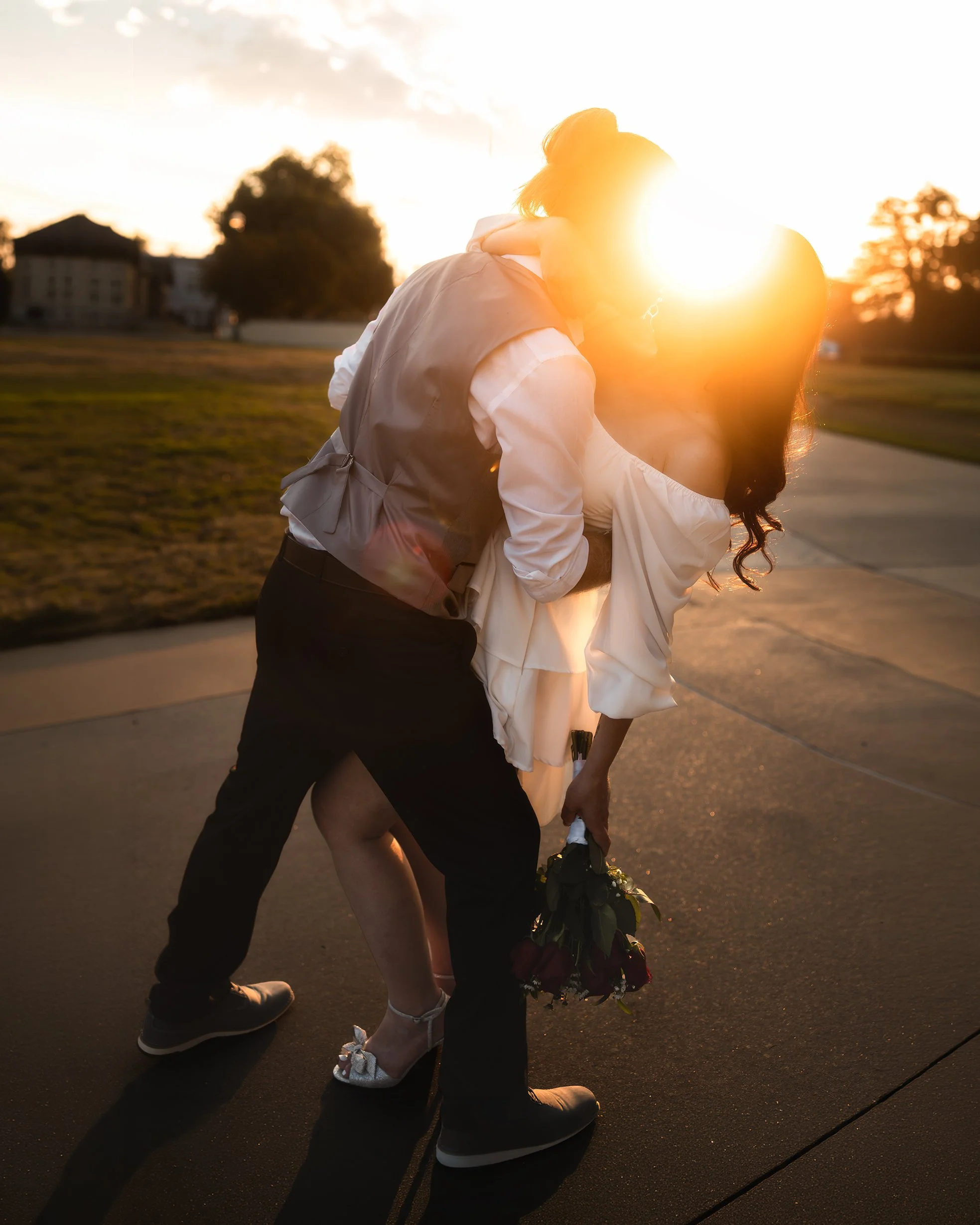 A couple embracing outdoors during sunset, with the sun behind them creating a warm glow, and one person holding flowers. Golden hour. newly weds. courthouse wedding photos. elopement photos. Colorado elopement photographer. 