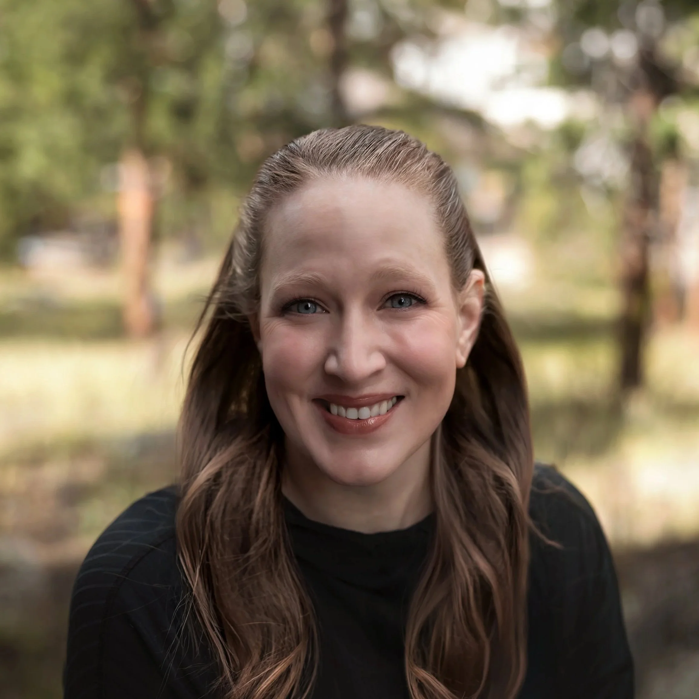 Close-up of a young woman's face with blue eyes, smiling, with a blurred background of trees and outdoors. Brenna Wakefield. Vow to Alpenglow Photography by Brenna Wakefield. Colorado elopement and wedding photographer.