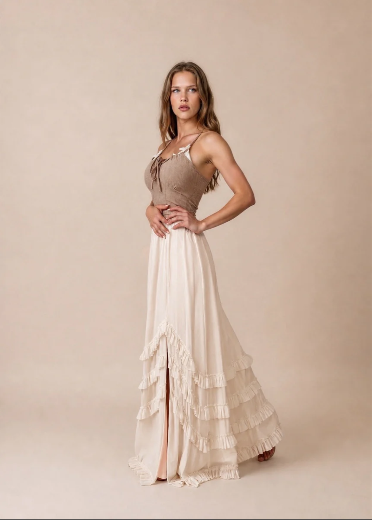 A young woman with long wavy hair wearing a beige and cream dress with ruffles, standing against a plain beige background.