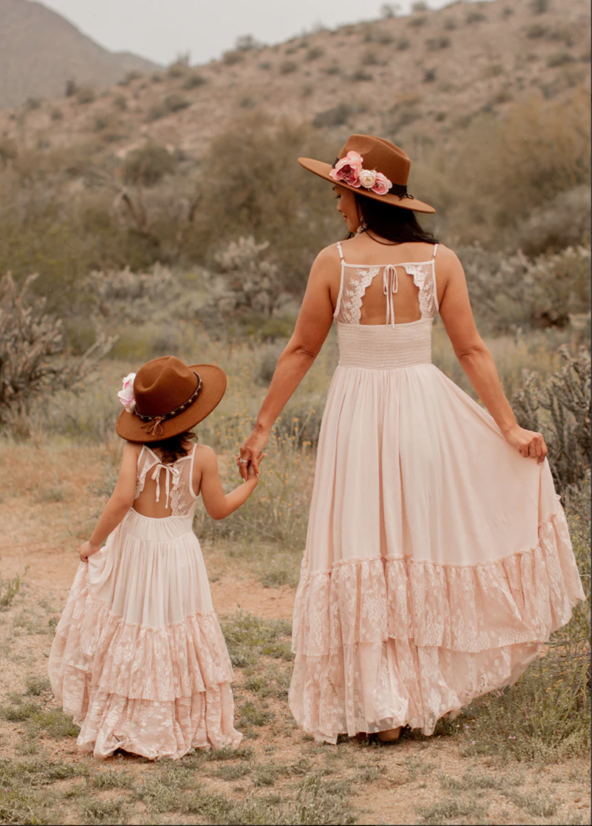A woman and a young girl hold hands while walking through a desert landscape. They are wearing matching long, flowy, cream-colored dresses with lace details and wide-brimmed hats decorated with pink flowers. The background shows desert vegetation and mountains.