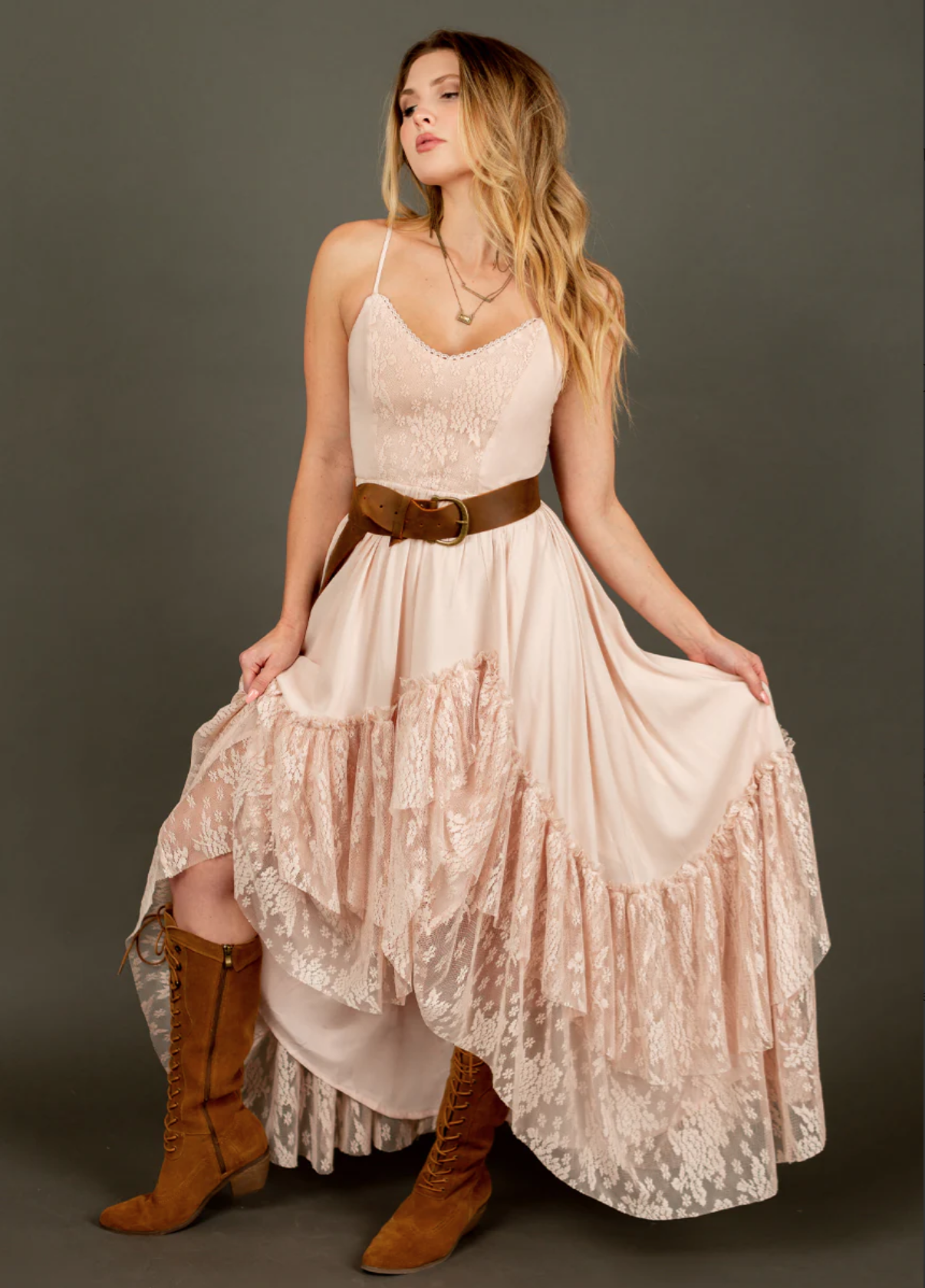 A woman with blonde hair wearing a beige lace and fabric dress, brown belt, and knee-high lace-up boots standing against a plain gray background.