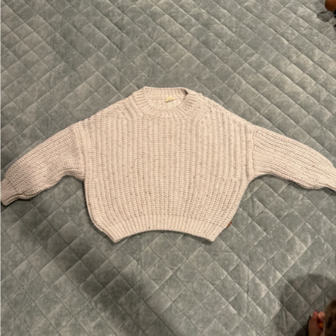 A beige, knitted cropped sweater with long sleeves laid flat on a gray quilted bedspread.