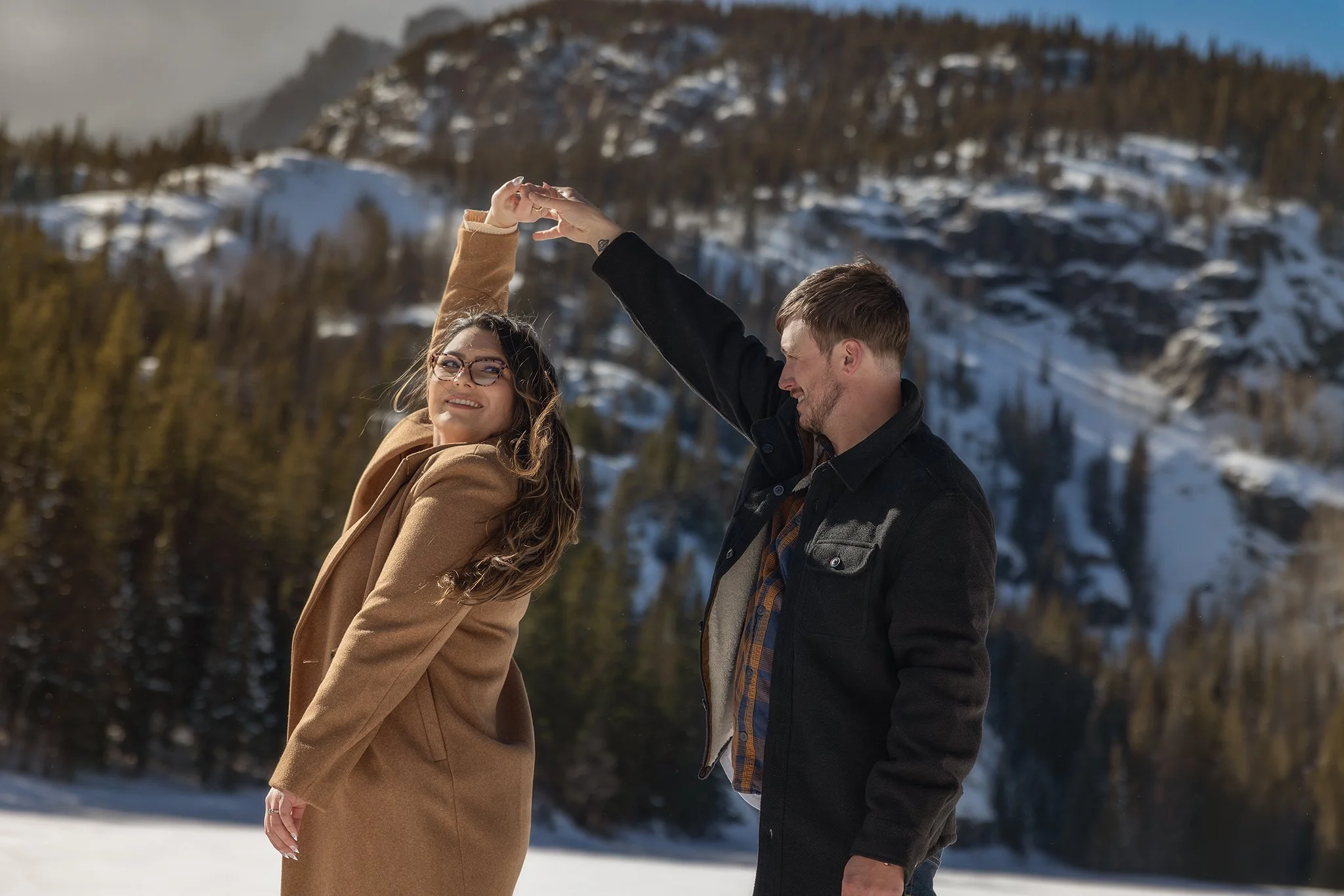 A couple holding hands and dancing outdoors in a snowy mountainous landscape with evergreen trees in the background. Bear lake. Colorado engagement photoshoot. Colorado engagement photographer.  Estes Park Colorado storytelling photographer. 