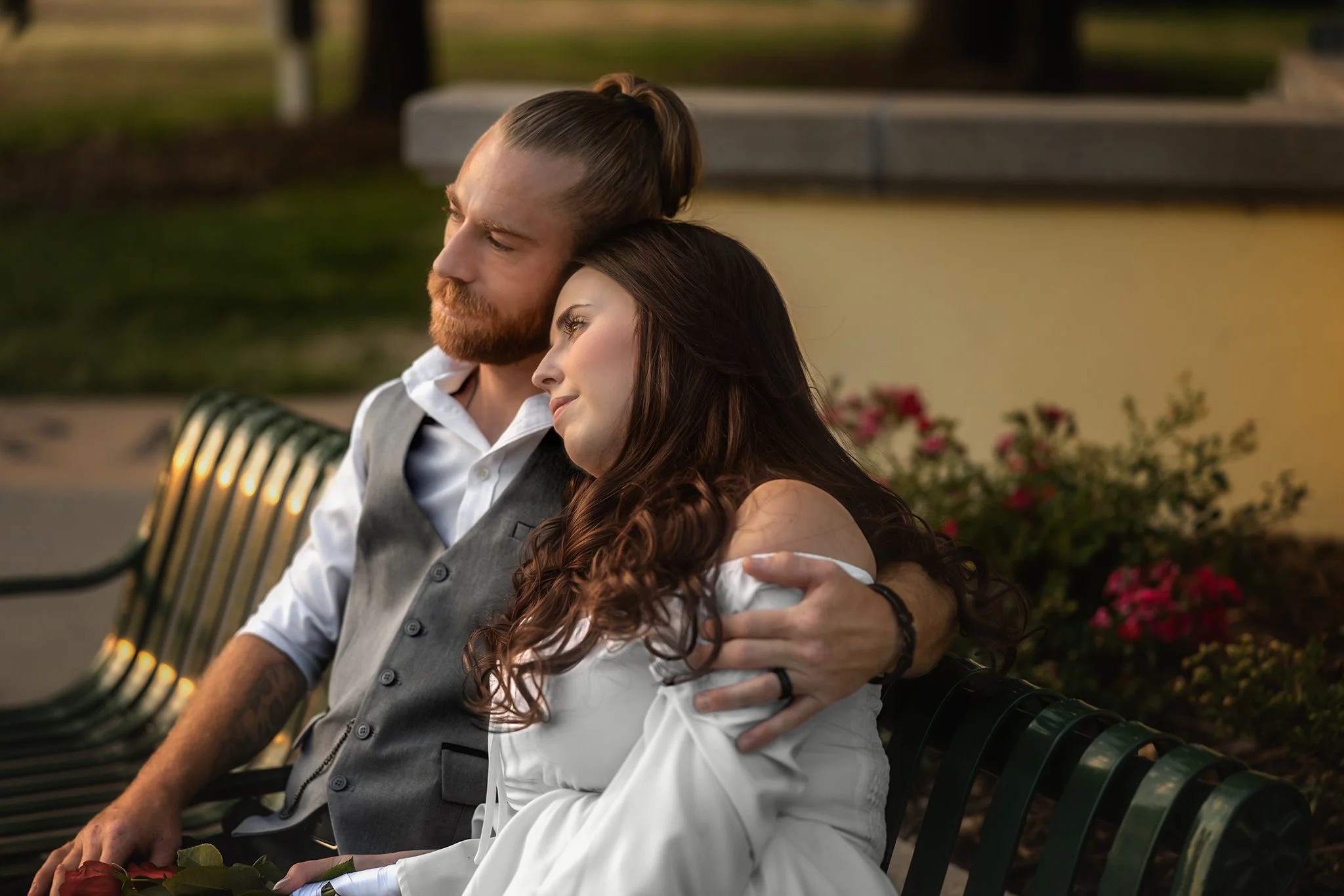 A man with red hair and beard in a gray vest hugging a woman with long dark hair in a white dress sitting on a park bench during sunset. Newly weds. Courthouse wedding. Greeley Colorado. Golden Hour