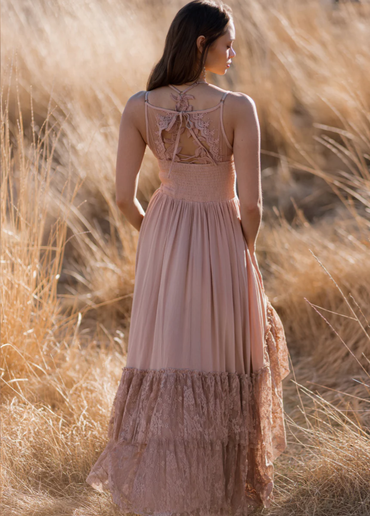 A woman in a long, pink lace dress with a lace back design, standing in a field of tall, golden grass, facing sideways.