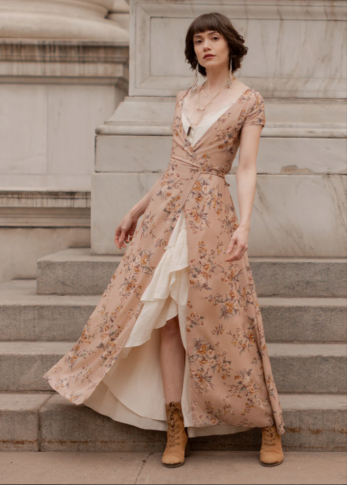 Woman standing on stairs wearing a floral dress with a slit, layered necklaces, earrings, and tan lace-up boots, in front of a stone building.