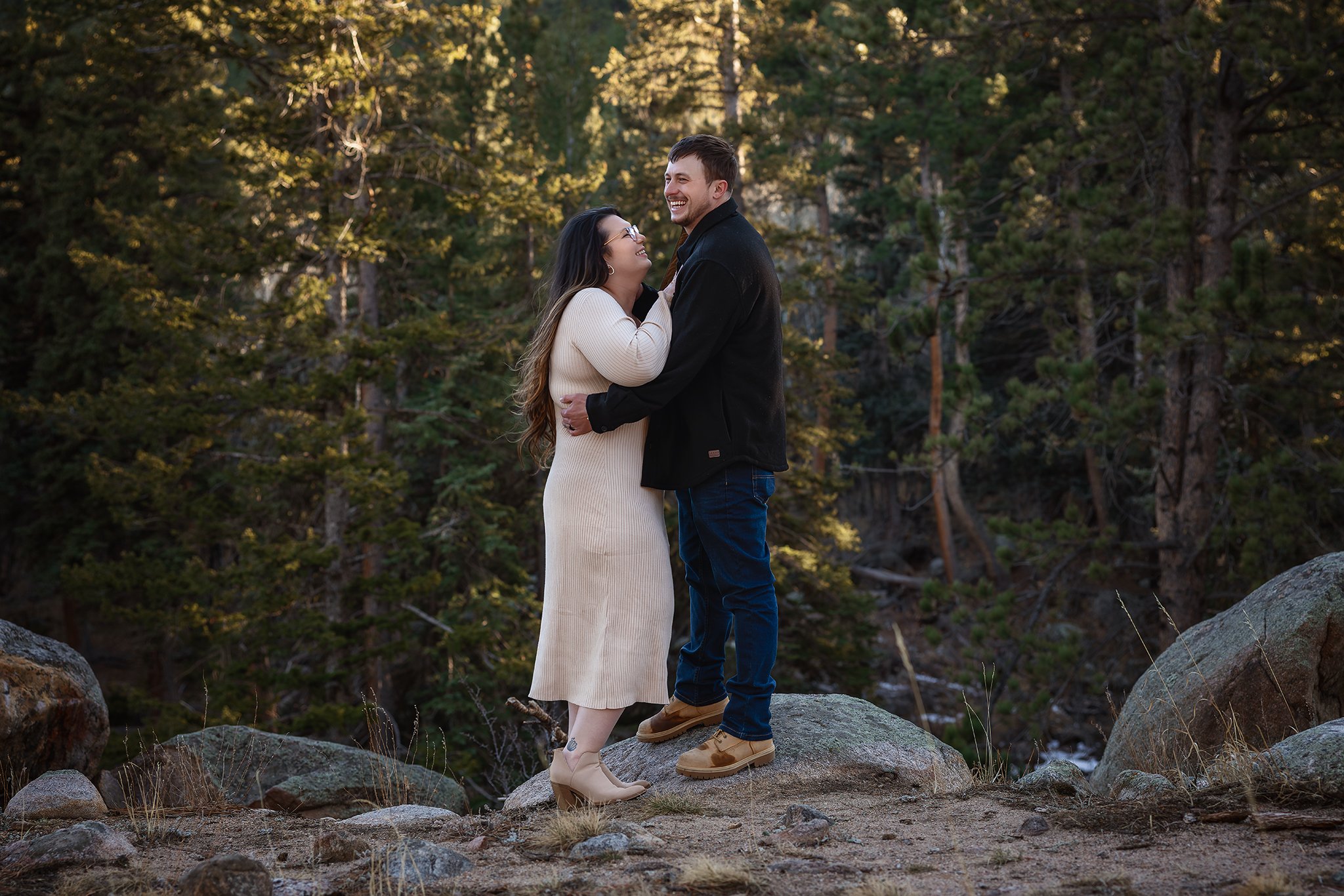 A couple standing on rocks in a forest, smiling and looking at each other.