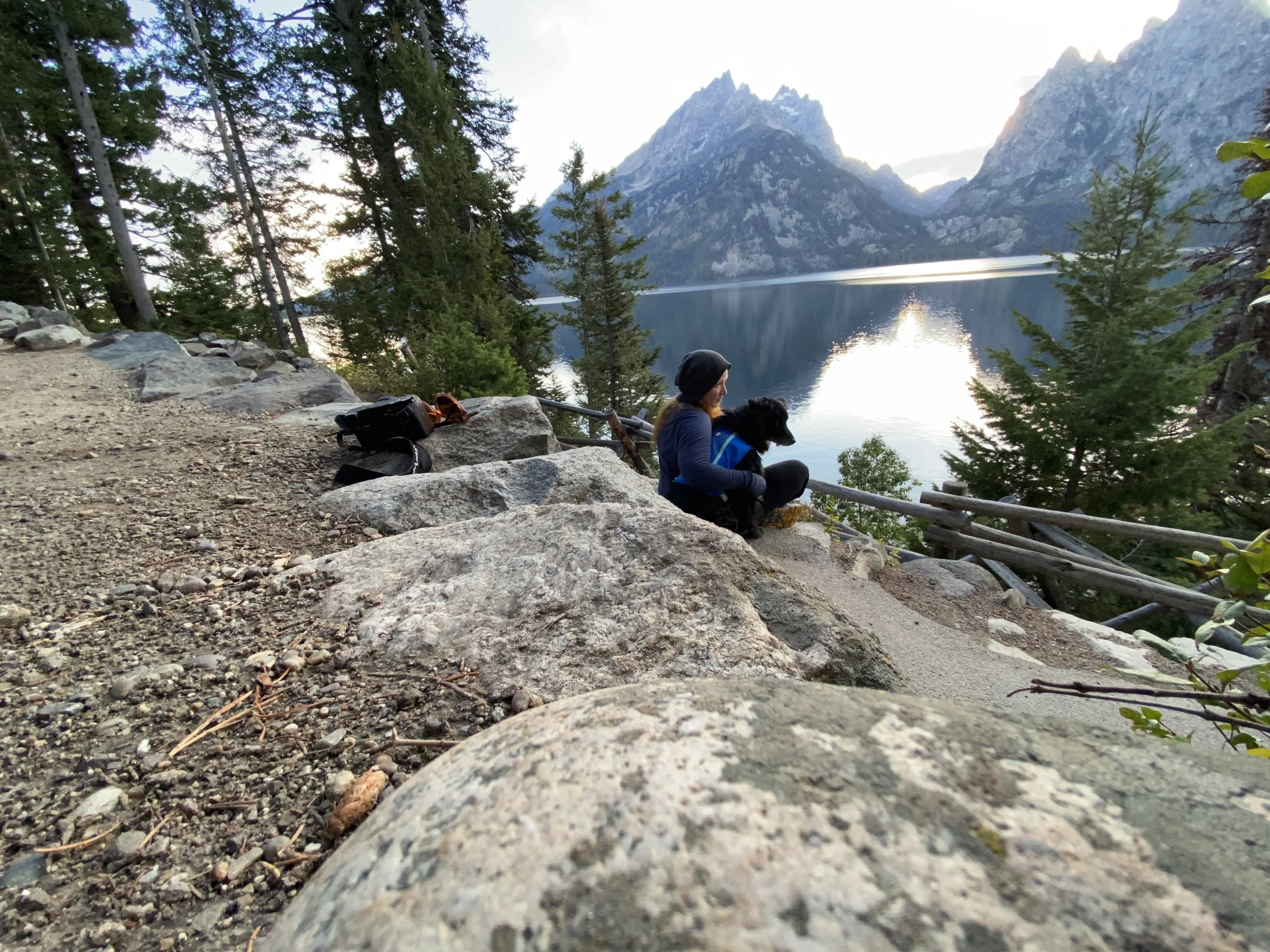 A woman with a dog sitting on rocks near a lake with mountains in the background, surrounded by trees, during sunset.