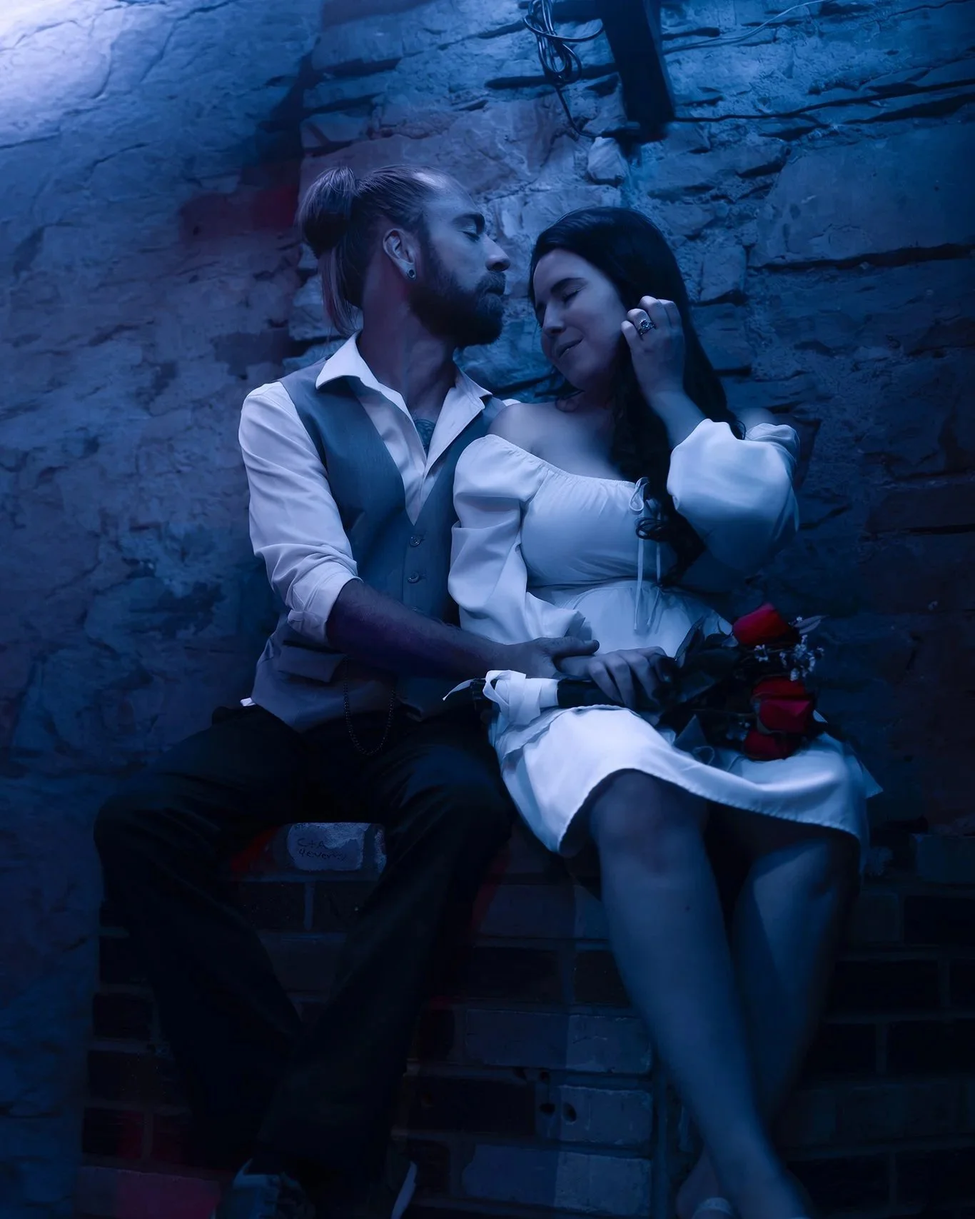 A couple sitting closely together against a rocky wall, with a bouquet of red roses on their lap, in a dimly lit setting with blue lighting. Newly weds sharing an intimate moment after the vows in their favorite arcade. Greeley Colorado arcade.