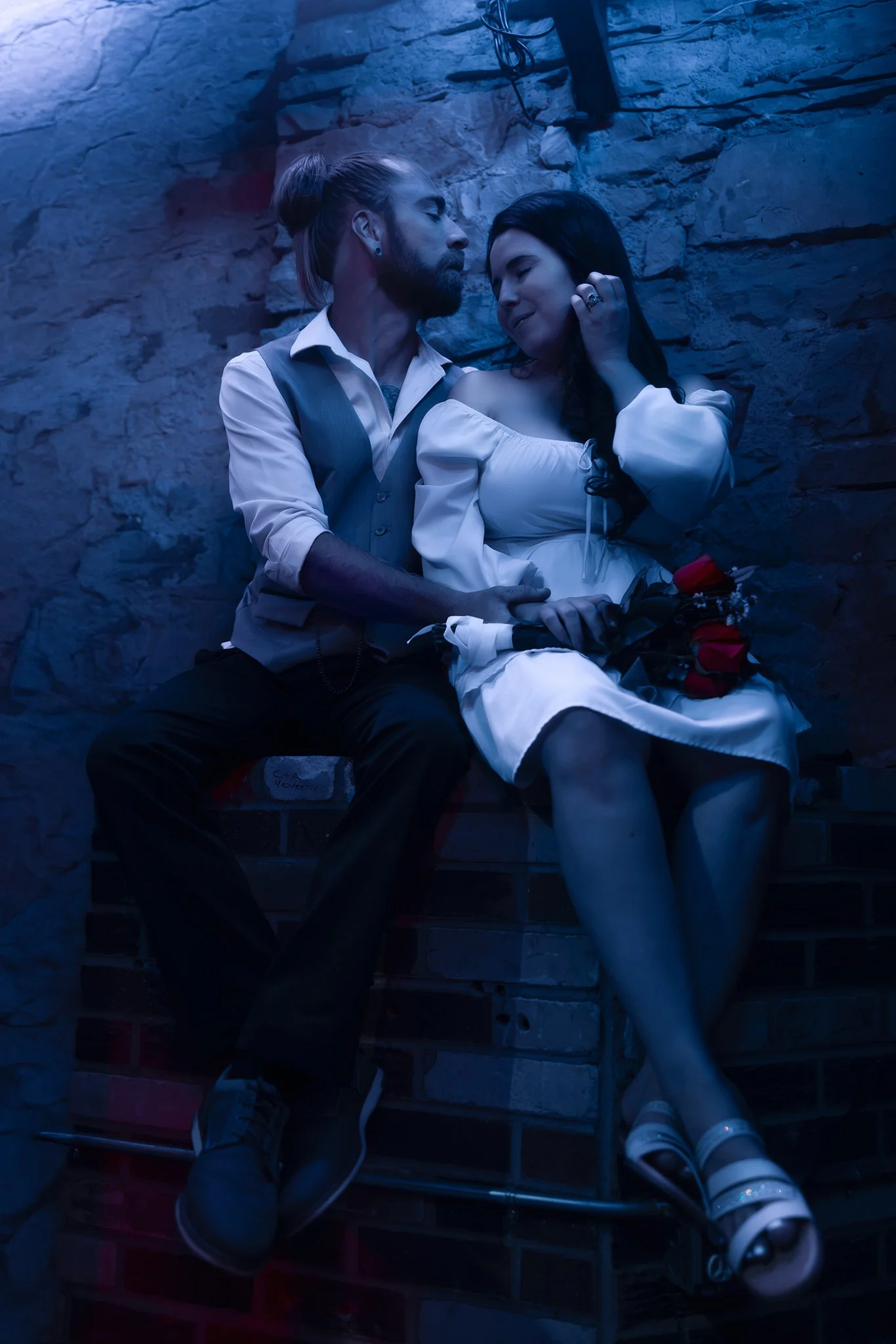 A man and woman sit close together on a bench against a stone wall, with the man holding a bouquet of red roses, both smiling softly. Newly weds. Courthouse Elopement. First date in arcade. Colorado elopement photoshoot. 
