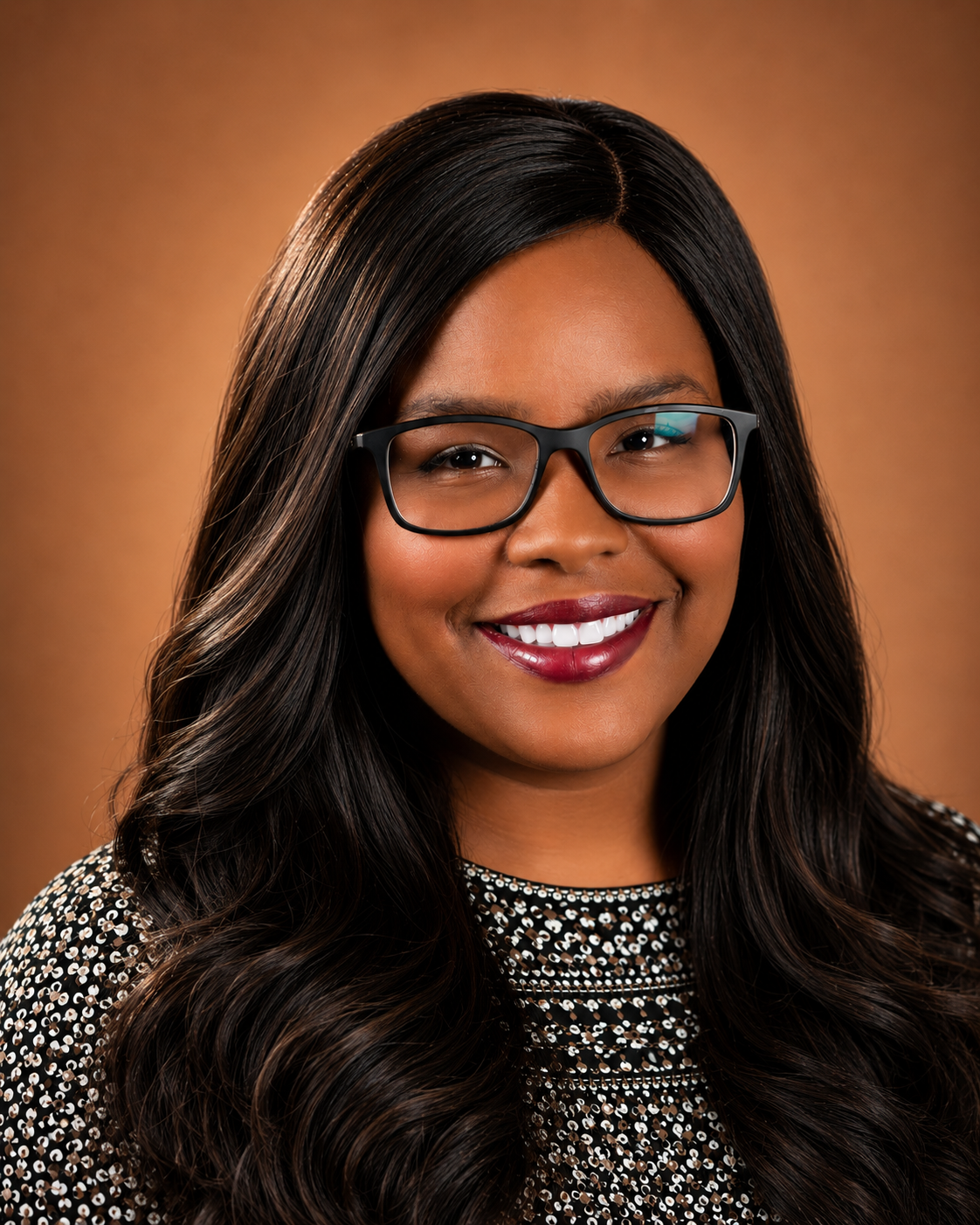 Portrait of a smiling woman with long dark hair, wearing glasses and a patterned black and white top, against a warm brown background.
