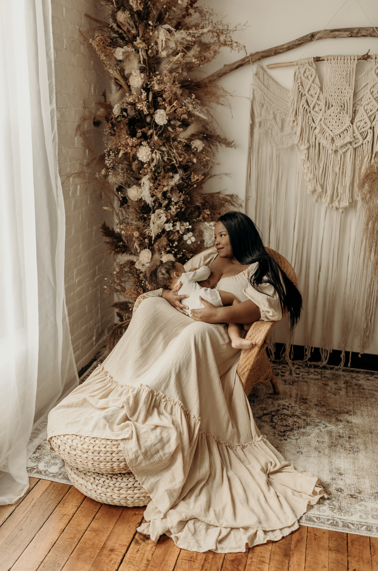 A woman with long black hair wearing a beige dress with puff sleeves sitting in a wicker chair, breastfeeding a baby. Behind her is a decorative dried floral arrangement and a macrame wall hanging. The room has white brick walls, a window with white curtains, and a wooden floor with a patterned rug.