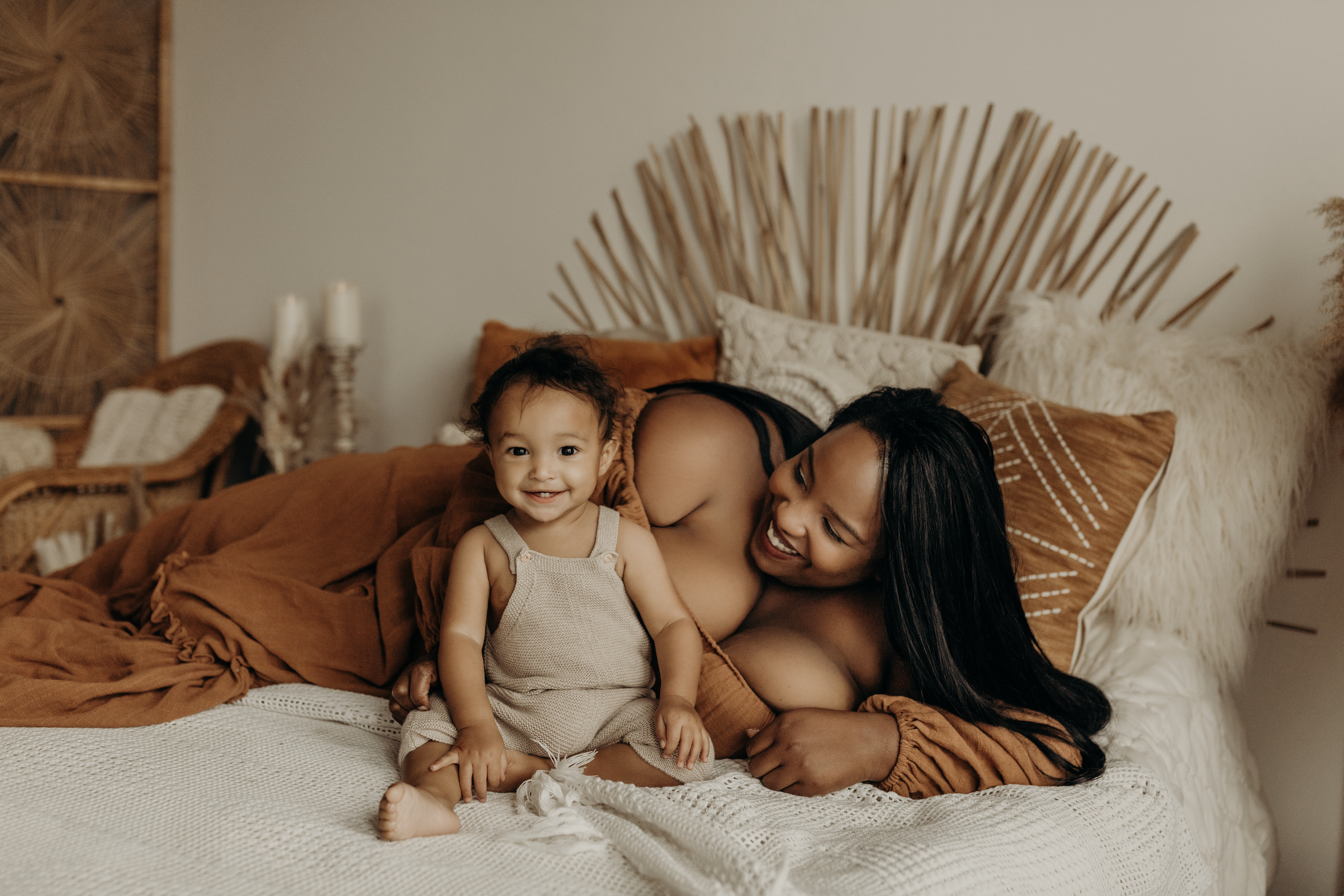 A woman and a young girl on a bed in a warmly decorated room, smiling at the camera. The woman is lying on her side, partially behind the girl, with dark hair and a friendly expression. The girl, sitting in front, has curly hair, a light-colored tank top, and a cheerful smile. The bed has rust-colored bedding and patterned pillows. Behind them, a decorative headboard with a fan-like design and textured wall art are visible.