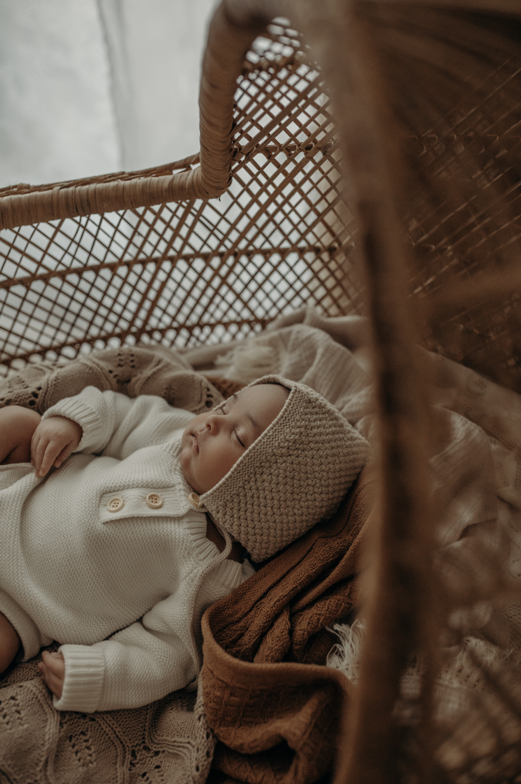 A sleeping infant in a cream-colored knit outfit and matching hat, lying on a blanket inside a wicker bassinet.