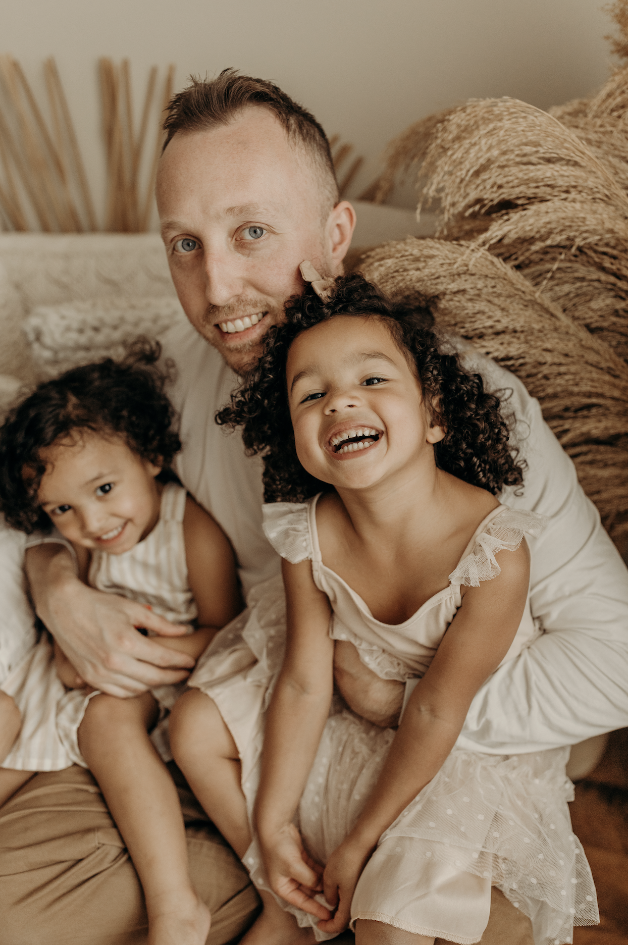 A man and two young girls sitting closely on a couch, smiling and looking at the camera, with neutral background and cozy decor.