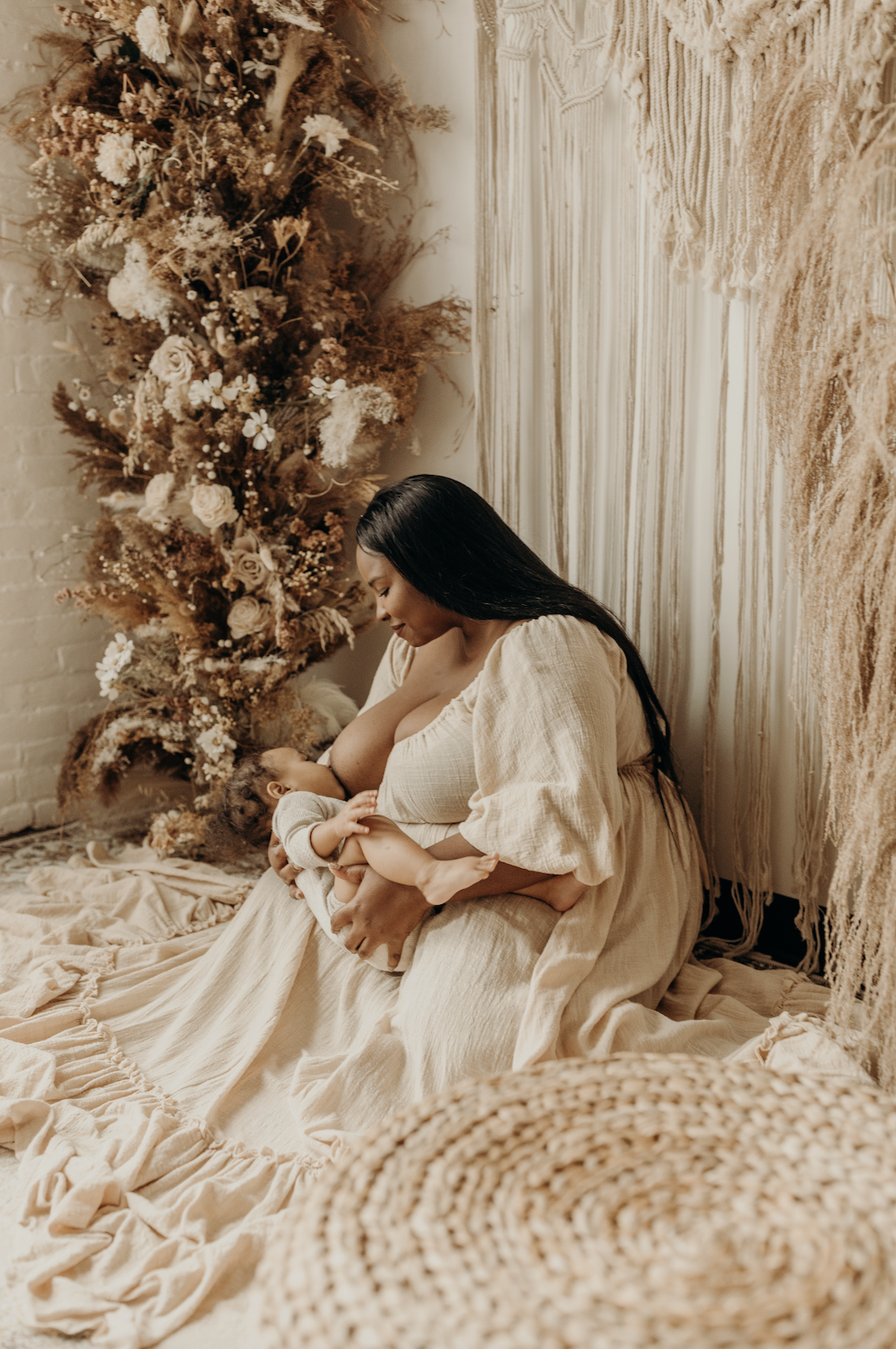 A woman in a beige dress sitting on a beige blanket on the floor, breastfeeding her baby. Behind her is a decorated Christmas tree with dried flowers and neutral-colored ornaments, and a textured wall hanging surrounds the scene.