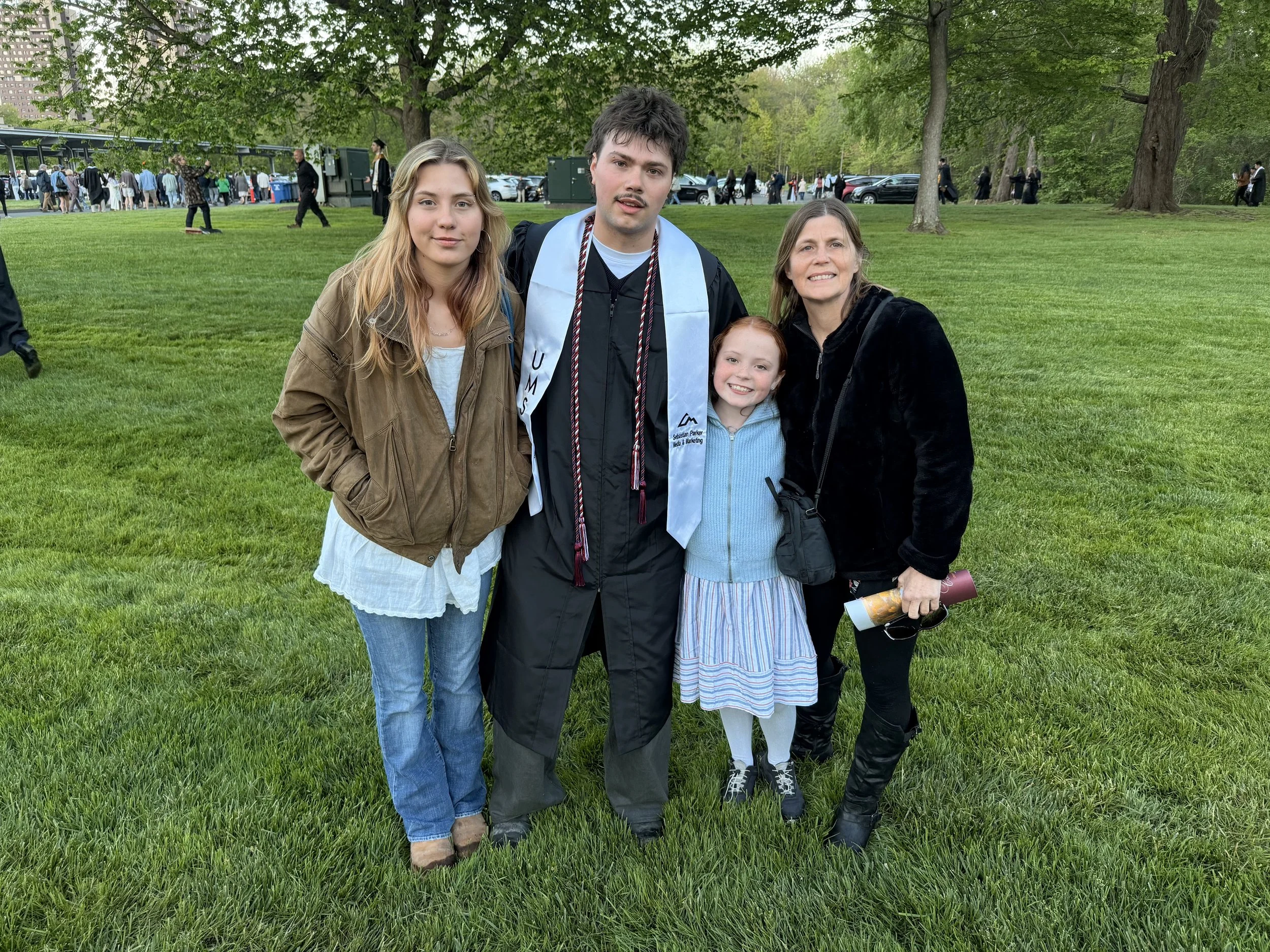 A group of five people, including a young man in graduation attire, two women, and two girls, standing on a grassy area in a park with many people in the background, trees, and parked cars. The young man is wearing a black graduation cap and gown with honor cords and a sash. The women and girls are dressed casually, smiling and posing for the photo.