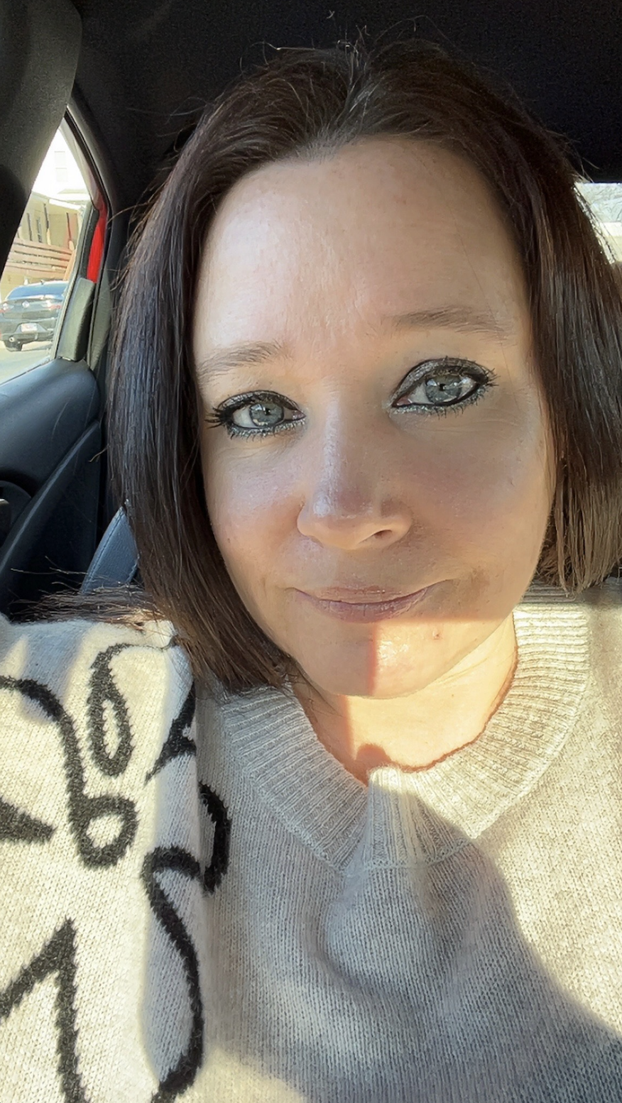 A woman with shoulder-length brown hair and blue eyes sitting in a car, wearing a light-colored sweater, with sunlight on her face.