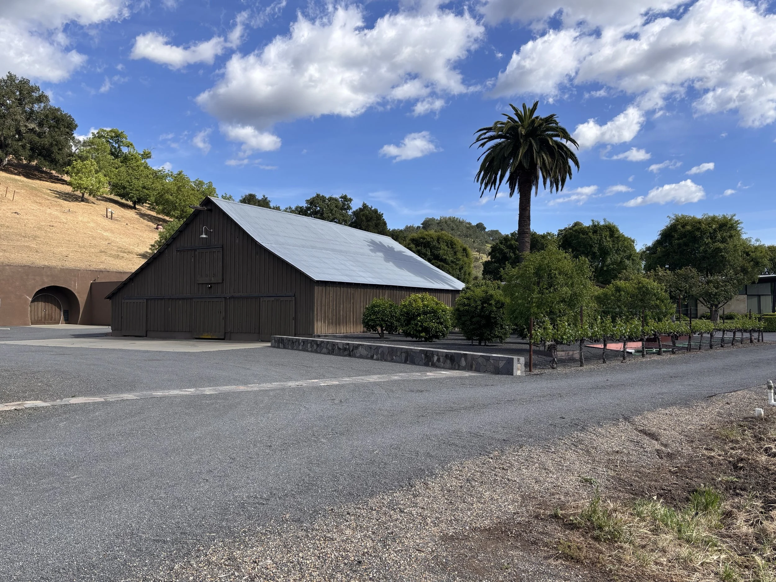 A large wooden barn with a metallic roof, set on a paved area with a gravel edge, surrounded by green trees, a hillside with dry grass, and a bright sky with clouds.