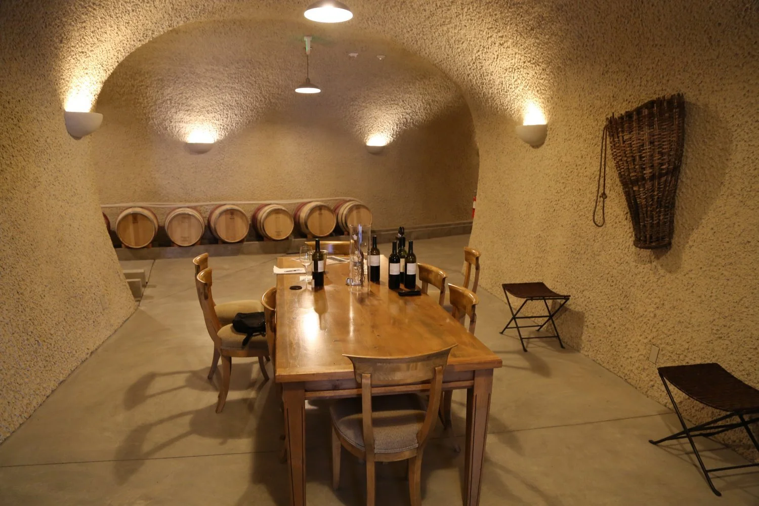 A wine cellar with a wooden table set for tasting, bottles and glasses on the table, wine barrels in the background, and textured beige walls with soft lighting.