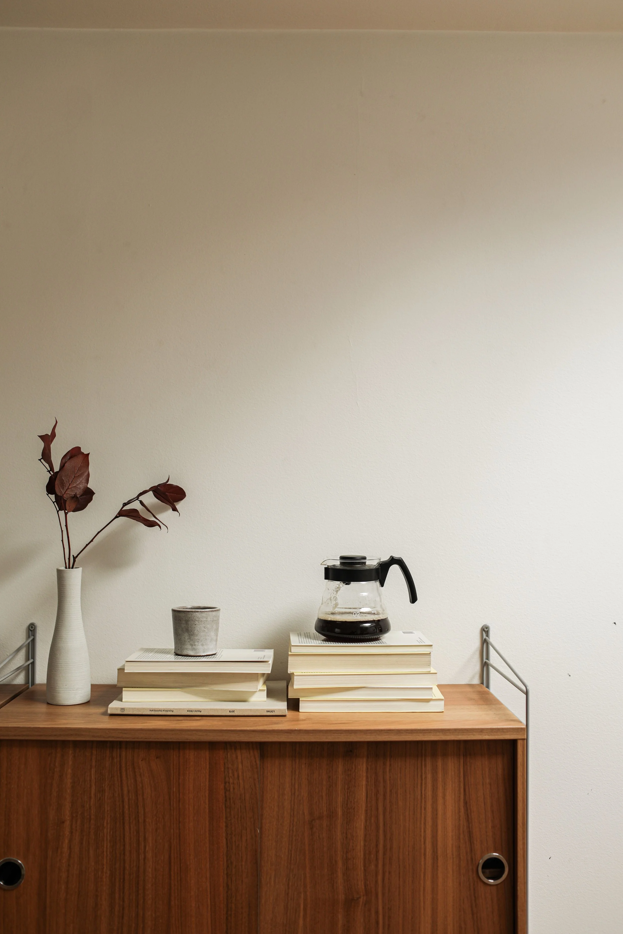 A wooden cabinet on a white table. On the table are stacked books, a coffee pot, a cup, and a vase with dried red leaves. The background is a plain off-white wall.