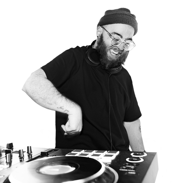 A male DJ with glasses, a beanie, and a beard, smiling and dancing while working at a DJ booth with turntables and a mixer.