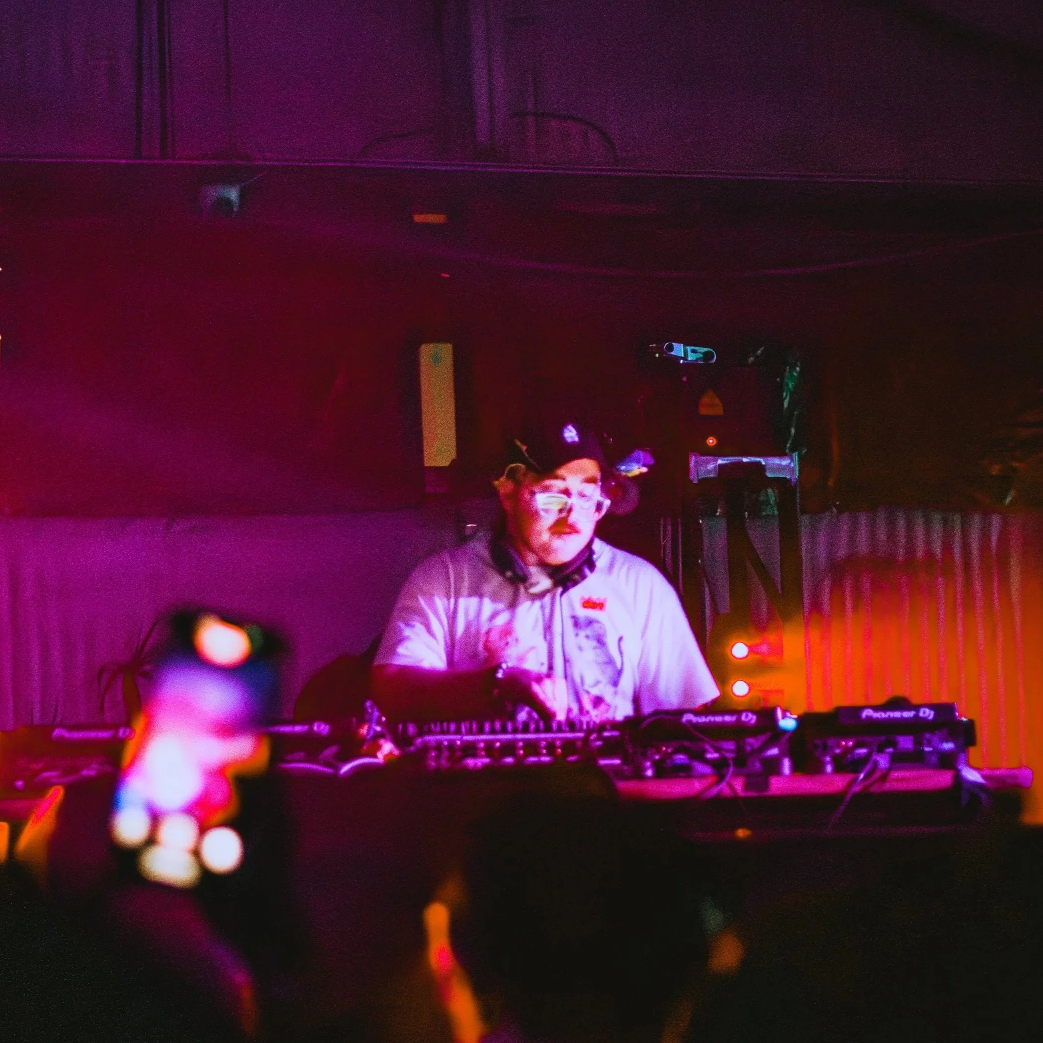 A person with glasses and a beard, dressed in a jacket, standing behind DJ equipment with colorful lights in a dark setting, likely at a party or nightclub.